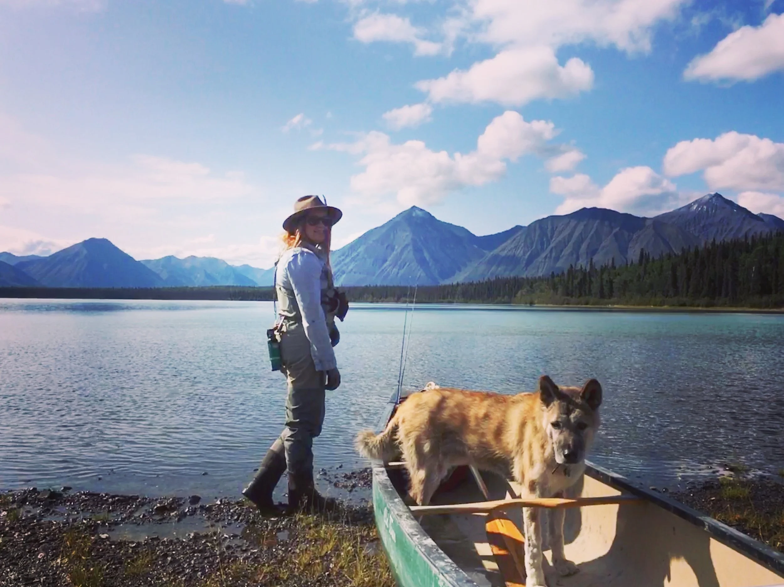 A woman with a hat and sunglasses standing by a lake with mountains in the background, accompanied by a large golden retriever dog.