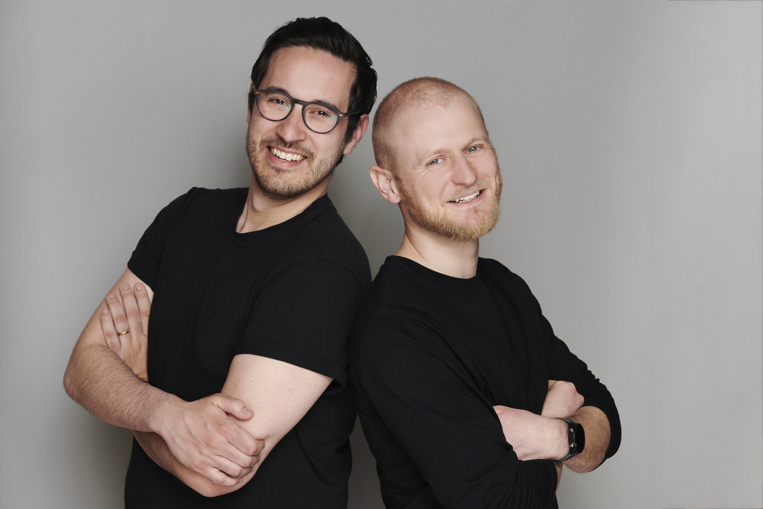 Two men standing back-to-back with arms crossed, smiling against a gray background.