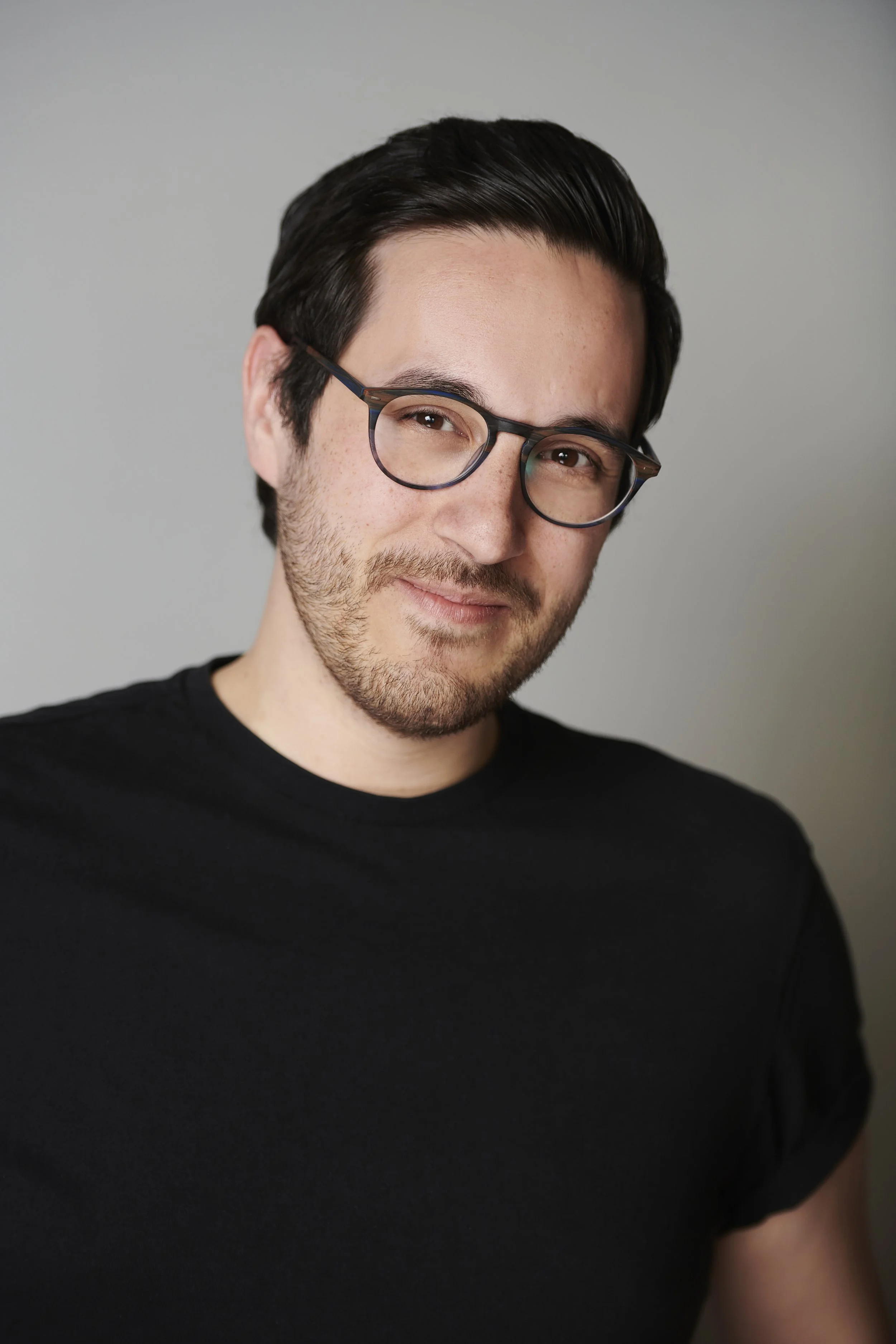 A young man with dark hair, glasses, and a light beard, wearing a black t-shirt, smiling slightly at the camera against a plain gray background.