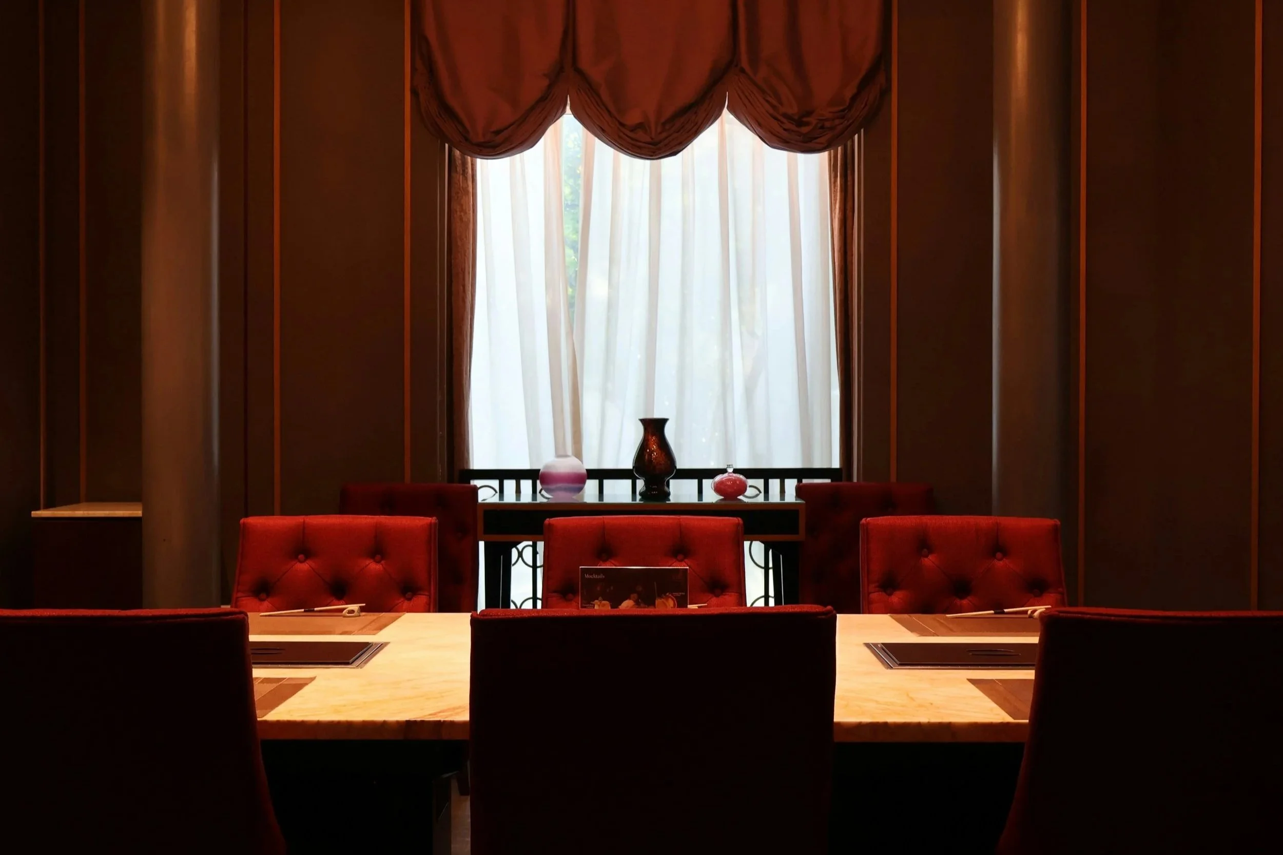 Private dining room with a long table, red upholstered chairs, and a softly lit window.