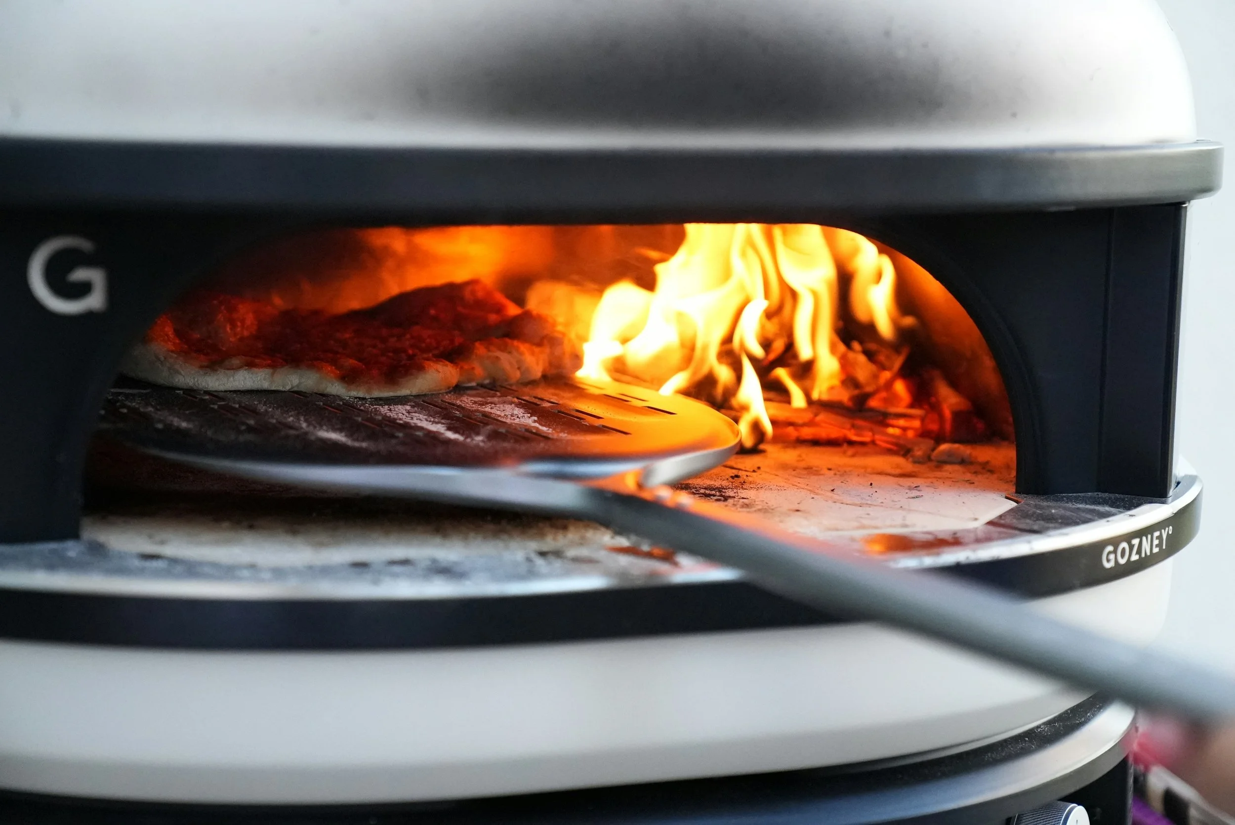 Pizza baking over an open flame in a portable wood-fired oven.