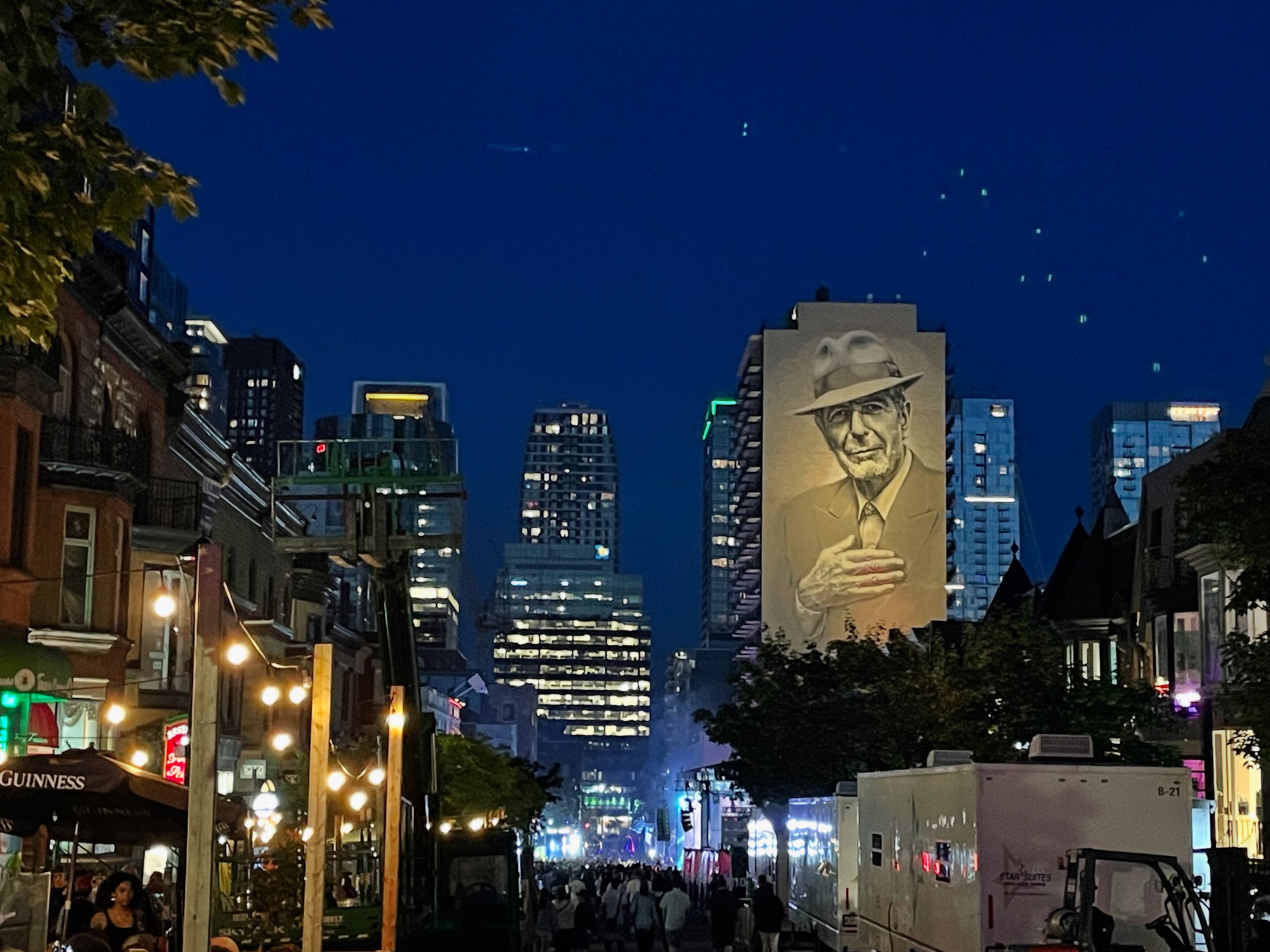 夜の都市の通りに広告の大きな絵画があり、中央には帽子をかぶった男性の顔が描かれている。周辺には街灯とビルの灯りが見える。
