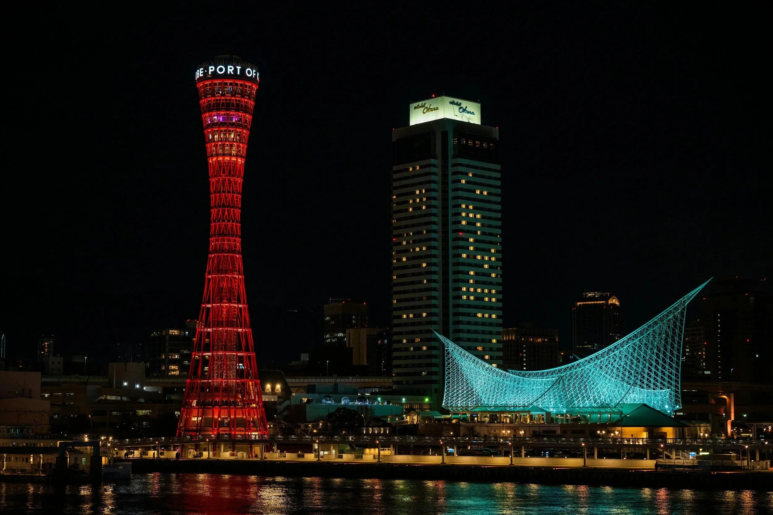 夜の港の風景。赤く照らされた展望タワーと、建物にはライトアップされた看板があり、橋もライトアップされている。