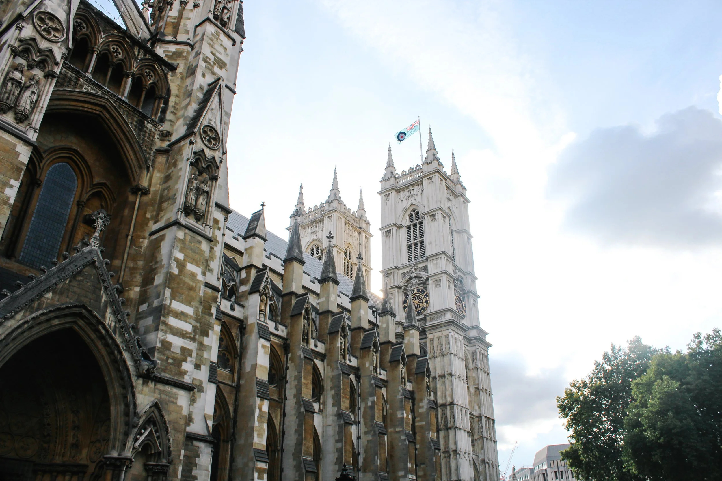 View of Westminster Abbey in London, England, with detailed stone architecture, tall towers, and flying buttresses, with a flag flying from one of the towers, cloudy sky in the background, and some trees on the right side.