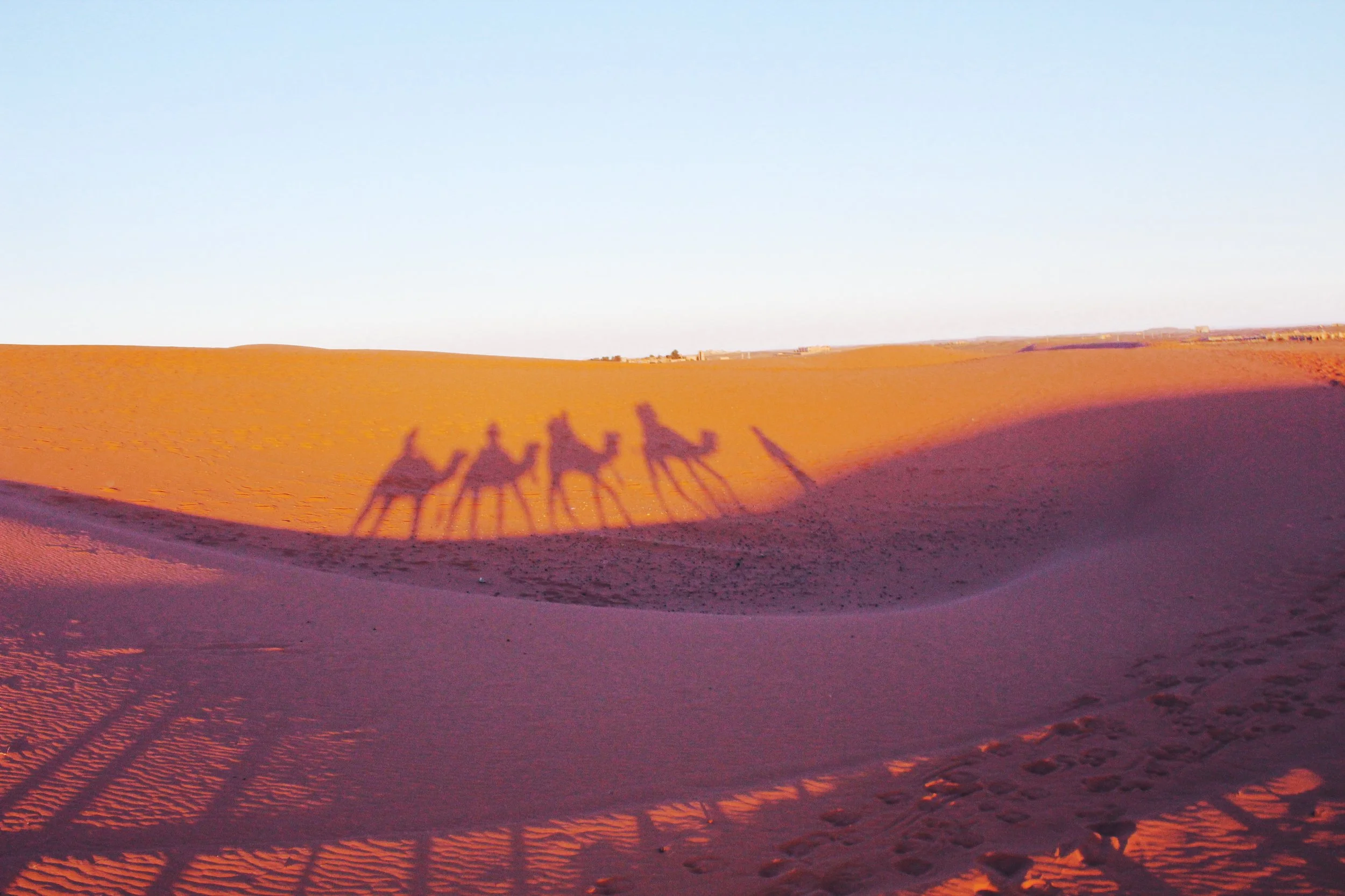 Shadows of three camels and their riders cast on sand dunes in Morocco at sunrise in a desert landscape.