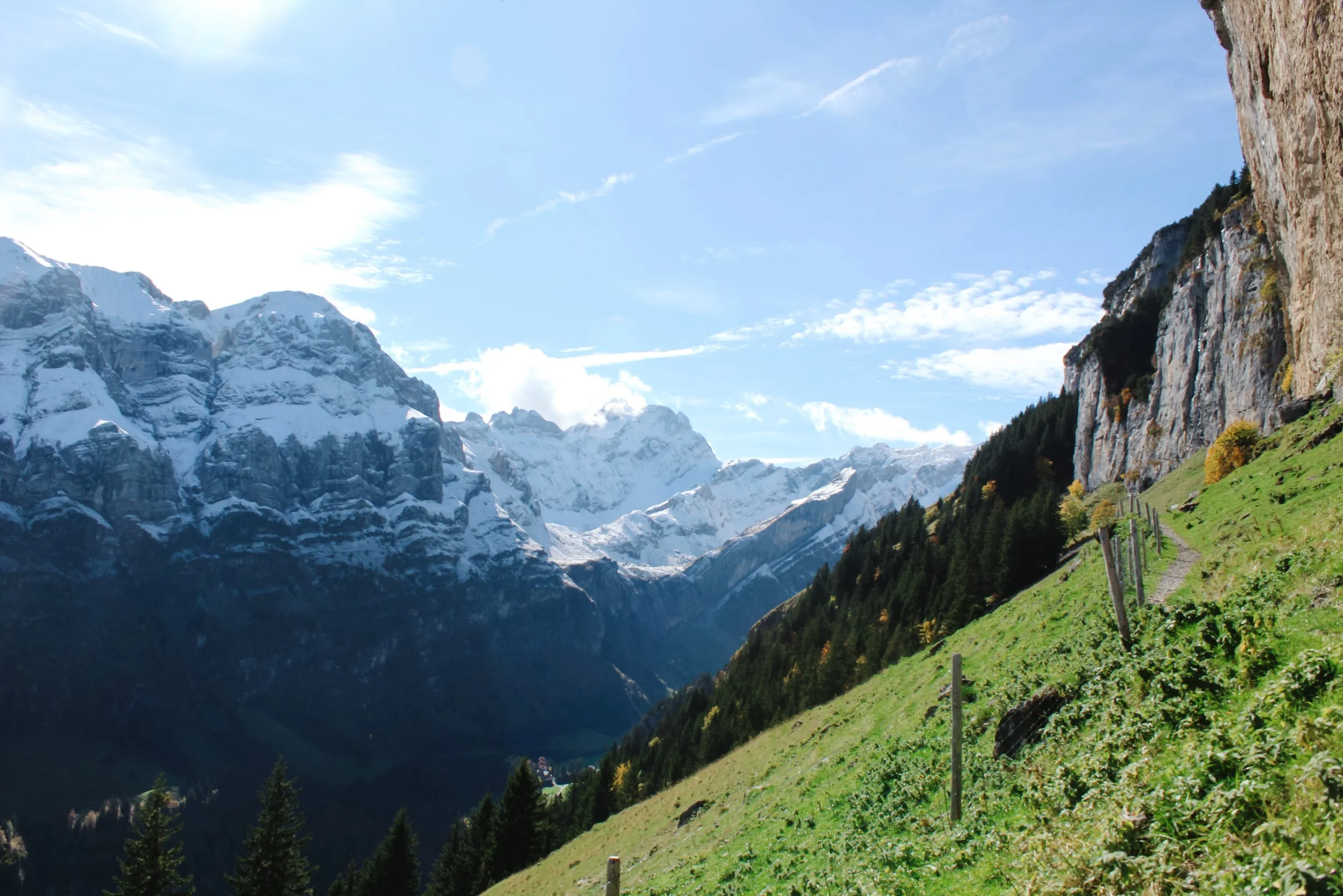 Snow-capped mountains in Ebenalp, Switzerland, under blue sky with some clouds, green hillside with trees, small trail, and a rocky cliff on the right.