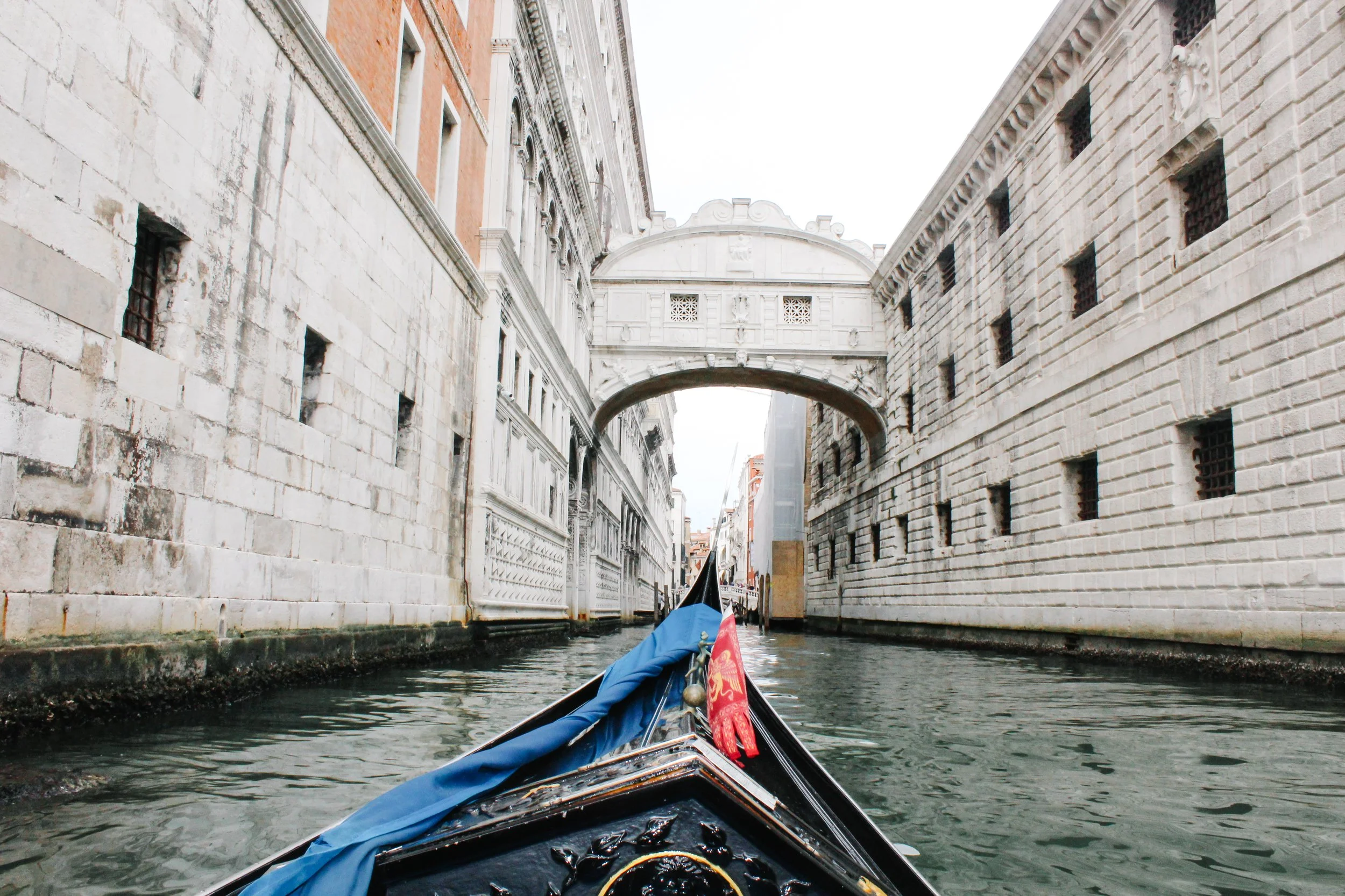View from a gondola on a canal in Venice, Italy, passing under a small arched bridge with historic stone buildings on either side.