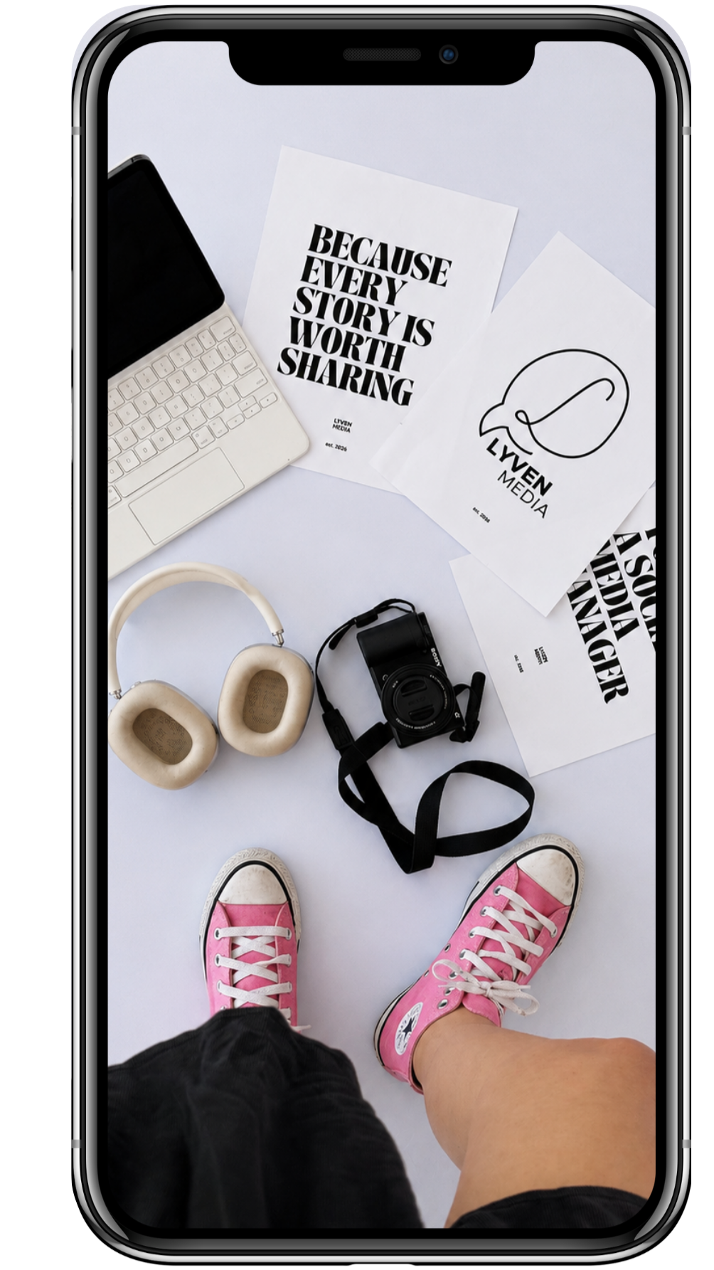 Photograph of a person wearing pink sneakers and black shorts, standing on a white surface surrounded by a camera, headphones, paper, a keyboard, and other objects, appearing as a flat lay or overhead shot.