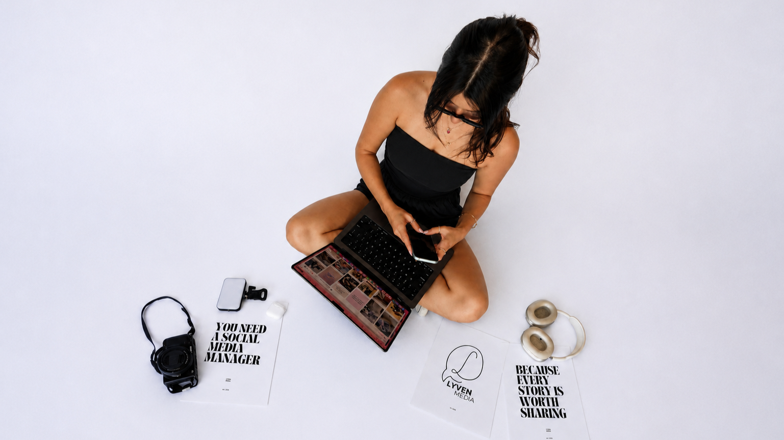 A woman sitting on the floor with various social media and photography-related items around her, using a tablet and smartphone.