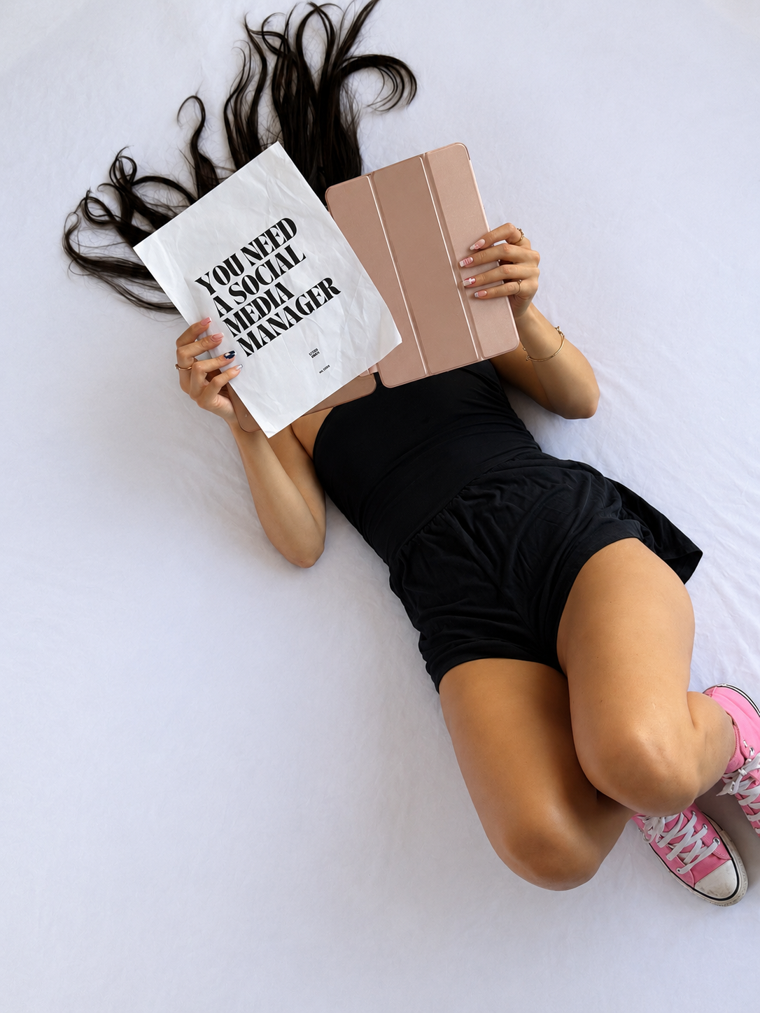 Person lying on a bed with long hair spread out, holding a tablet in one hand and a crumpled social media manager job offer in the other hand, wearing a black outfit, pink sneakers, and jewelry.