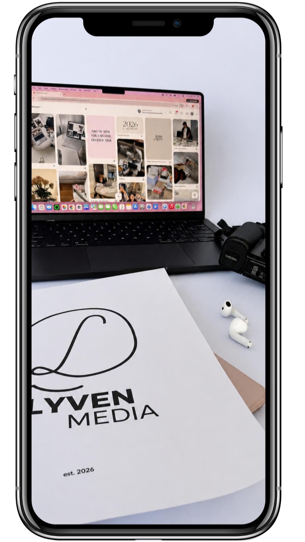 A smartphone screen showing a desk with a silver laptop displaying a Pinterest board, a white paper with 'LYVEN MEDIA' logo and 'est. 2026' in front of the laptop, a pair of white wireless earbuds, and a black camera charger.