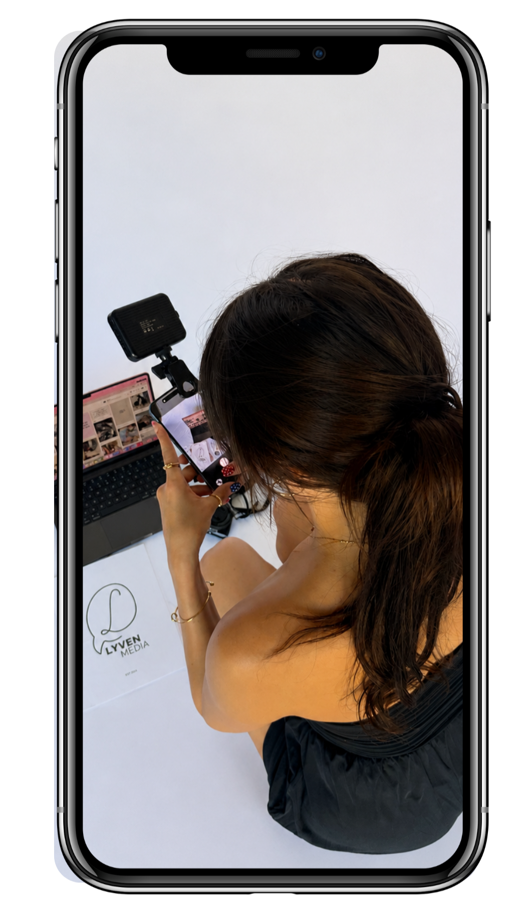 Woman with long dark hair taking a selfie with her phone, sitting at a table with a laptop and a logo printed on paper that says 'Liven Media'.
