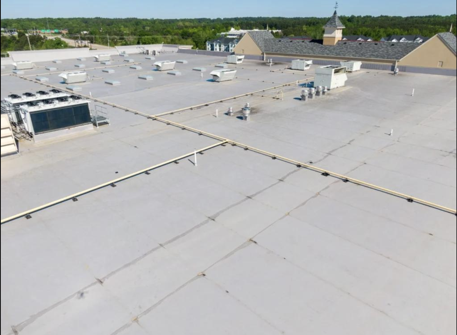 The flat rooftop of a commercial building with various HVAC units, vents, and piping systems, and houses in the background with trees and a blue sky.