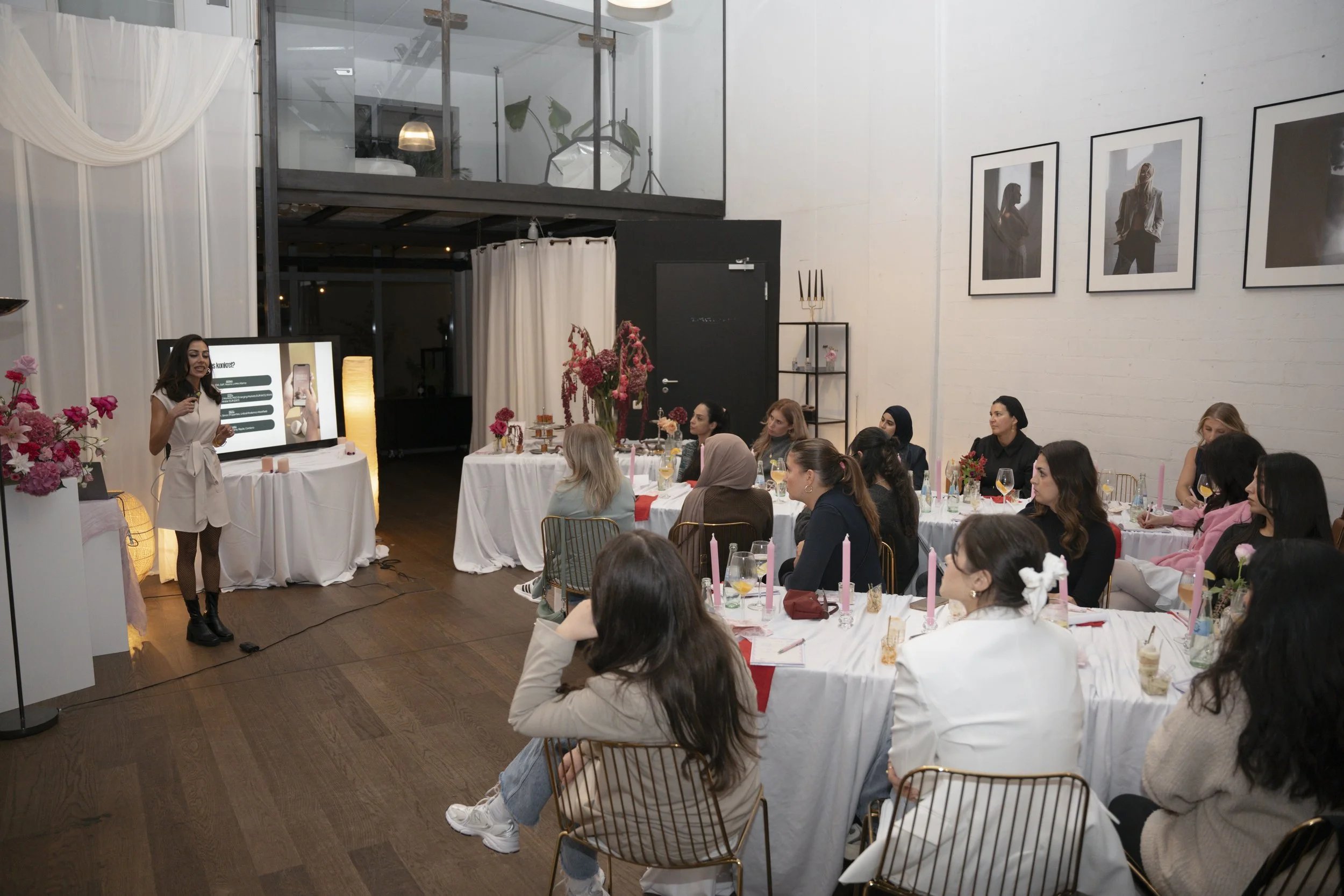 Anemone Paleko, Finance Mentorin, hält eine Präsentation vor Mitgliedern des Eve Tables bei einer Veranstaltung in einem modernen, hellen Raum mit weißen Wänden. Das Publikum sitzt an Tischen mit Kerzen und Blumen.