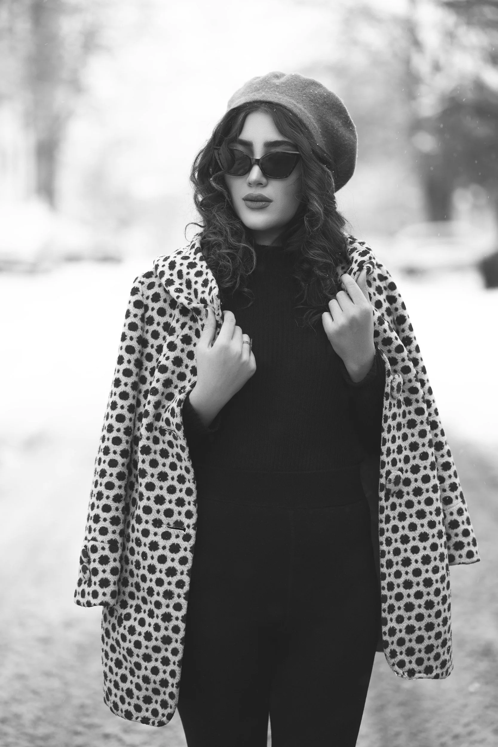 Black and white photo of a woman wearing sunglasses, a beret, a leopard print coat, and a black top, standing outdoors.