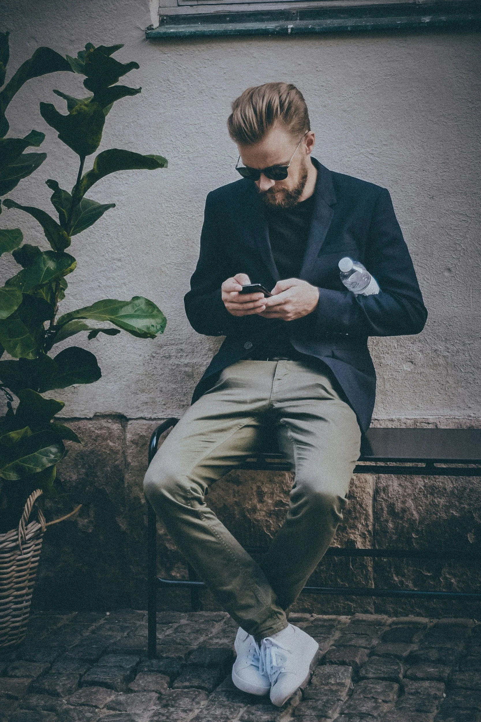 A man with blonde hair and sunglasses sitting on a bench, looking at his phone. He is wearing a black blazer, beige pants, and white sneakers, with a water bottle in his pocket. There are green plants and a textured wall in the background.