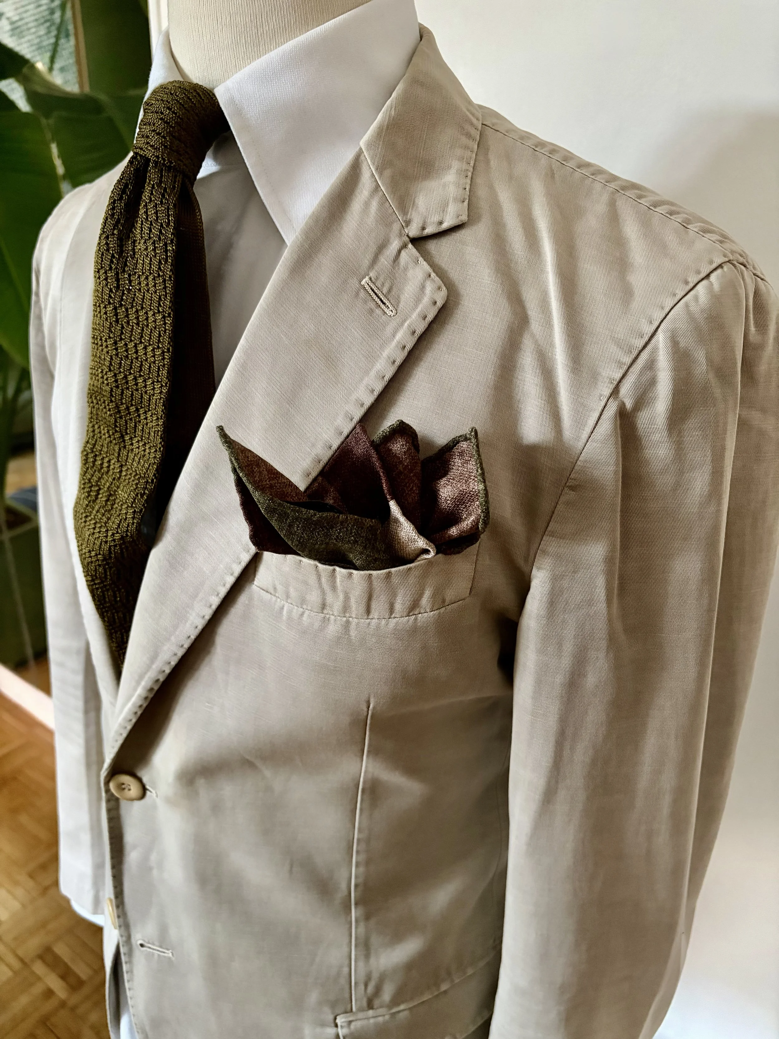 A beige blazer with a white shirt, a brown patterned tie, and a dark-colored pocket square were displayed on a mannequin.