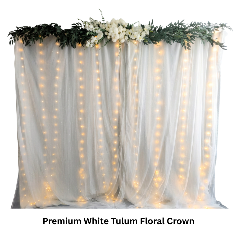 White tulle backdrop decorated with green foliage, white flowers, and hanging fairy lights, labeled as Premium White Tulum Floral Crown for a photo booth