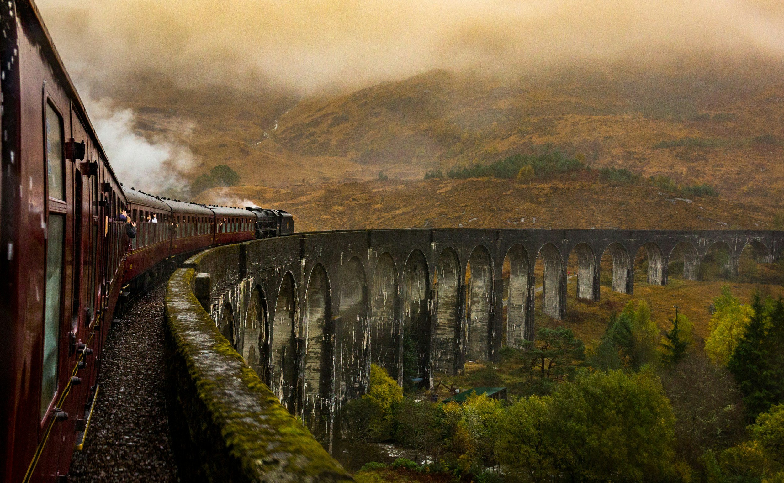 A vintage train with maroon carriages traveling on a curved viaduct through a foggy, green, mountainous landscape.