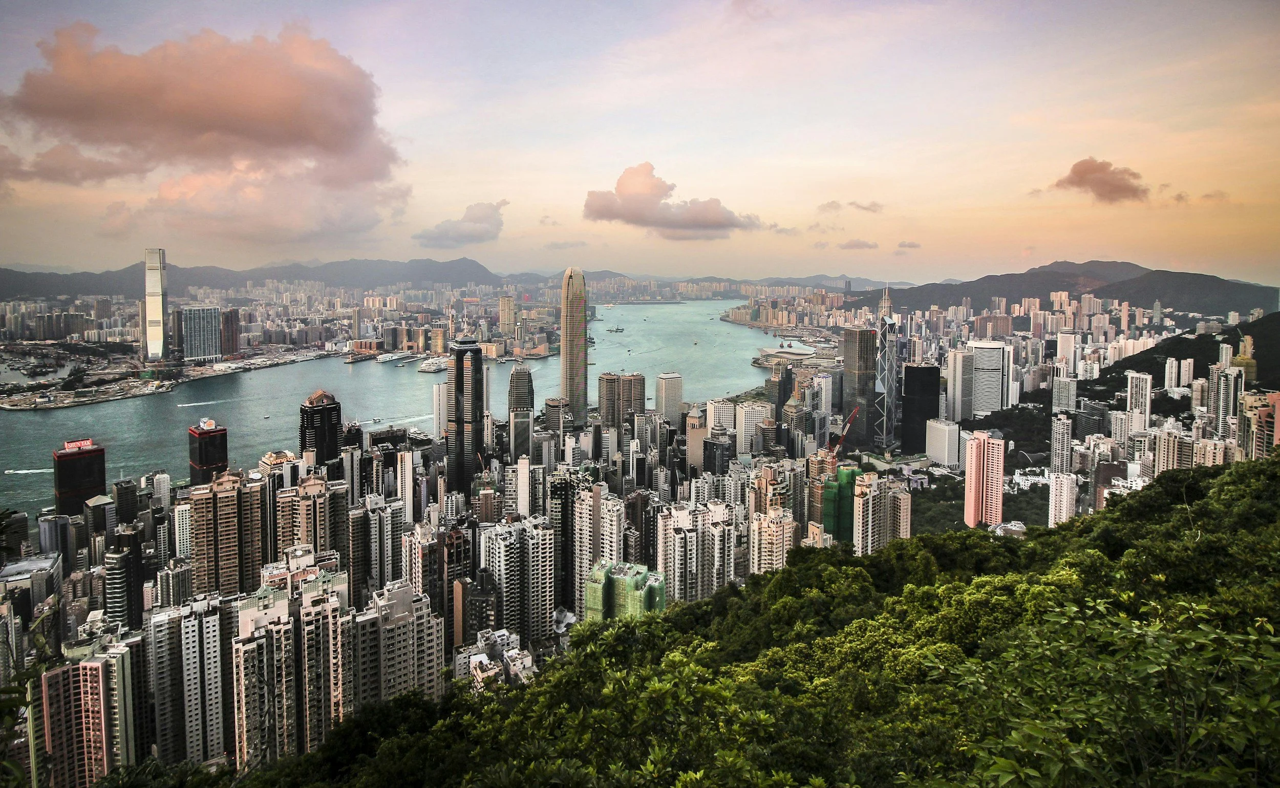 A city skyline during sunset with numerous high-rise buildings along a waterfront, mountains in the background, and a cloudy sky with pink and orange hues.