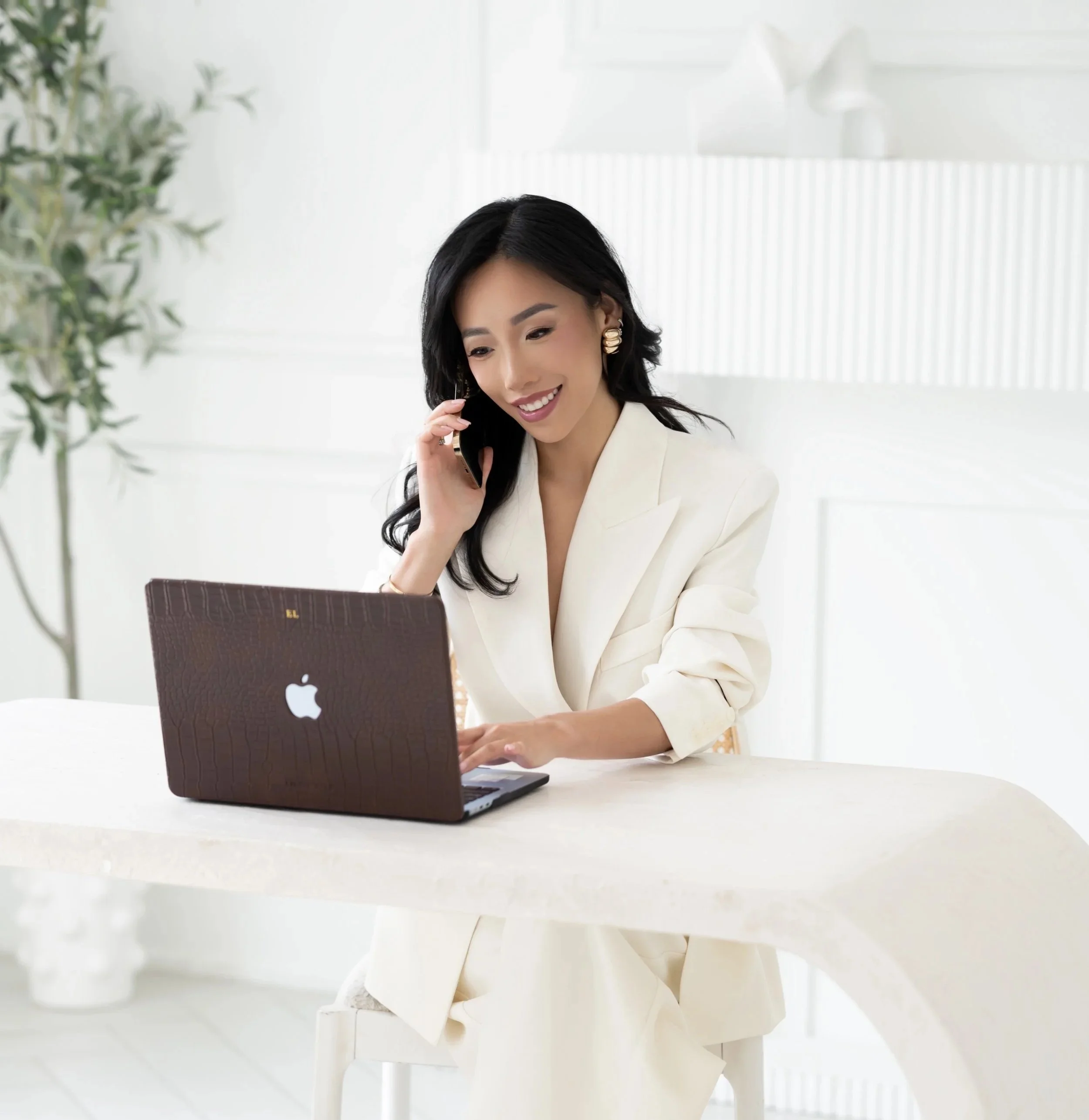 A woman with long dark hair wearing a white blazer, sitting at a white table in front of a laptop, smiling and talking on the phone in a bright, modern office with a plant and white decor.