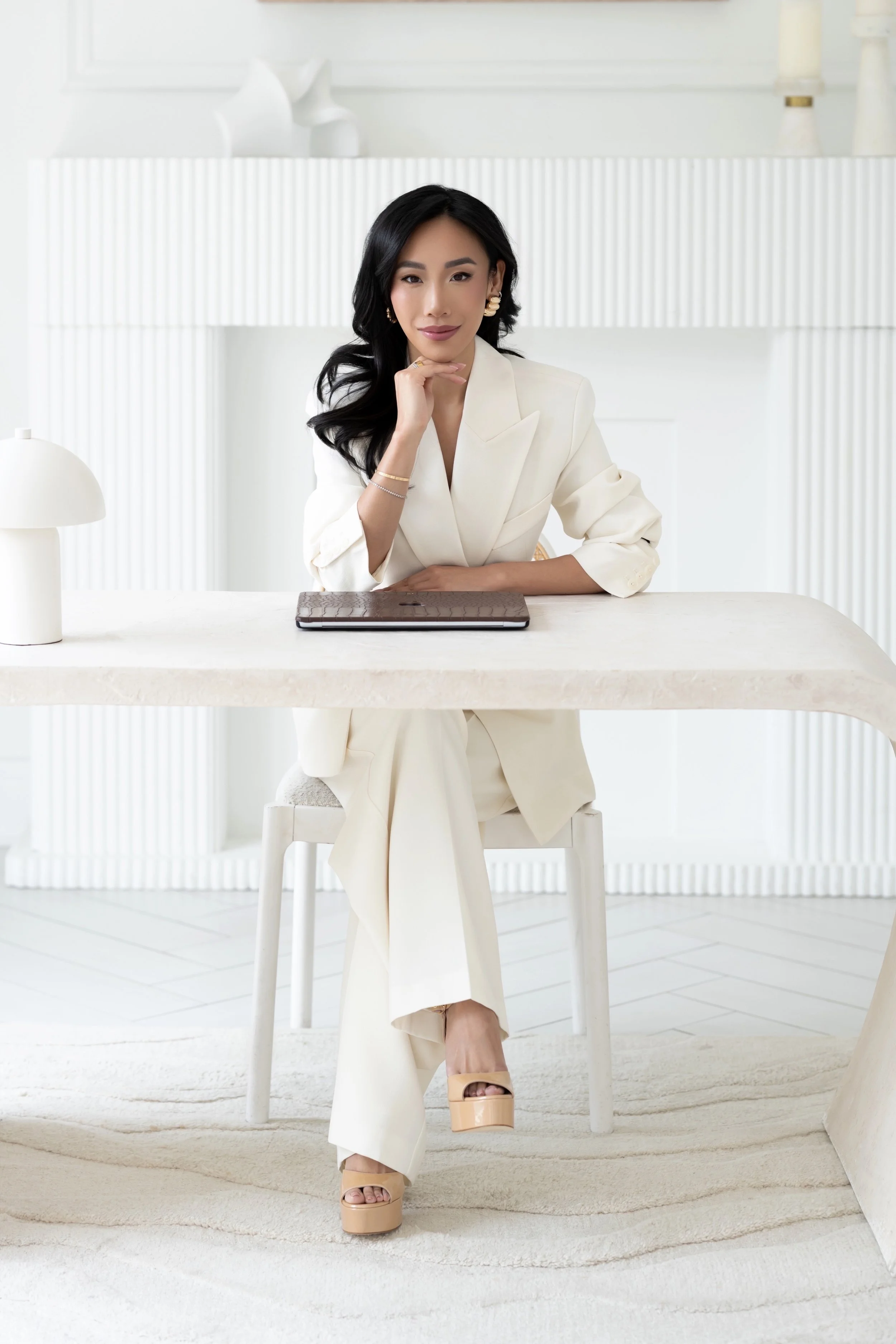 A woman with black hair wearing a white suit, beige platform heels, and gold jewelry, sitting at a white table in a bright minimalistic room.