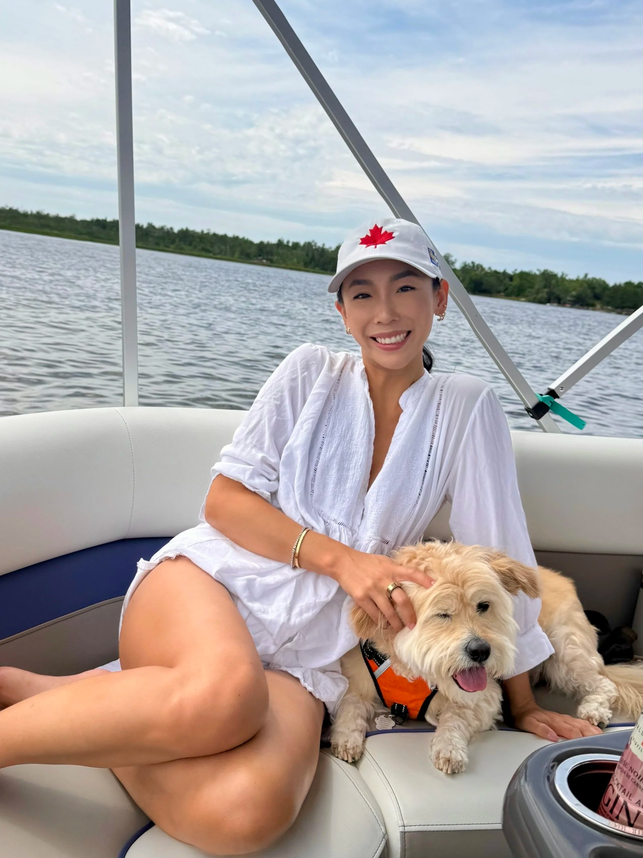 A smiling woman wearing a white cap with a red maple leaf and earrings, sitting on a boat with a yellow retriever dog. The dog is wearing an orange harness and appears happy. The background shows a body of water and a partly cloudy sky.