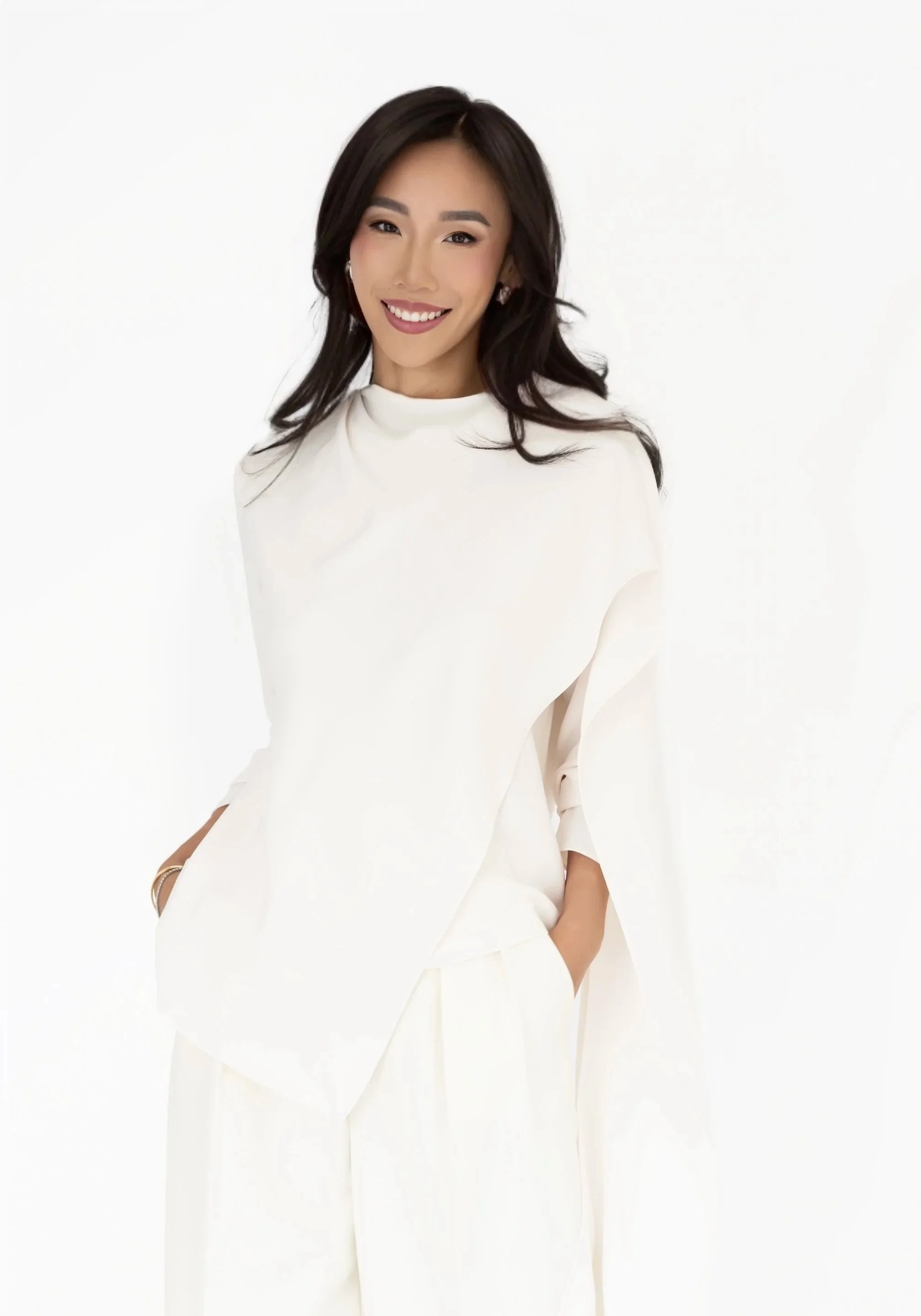 A woman with dark hair in loose waves, wearing a white top, smiling and winking at the camera, with a minimal white background.