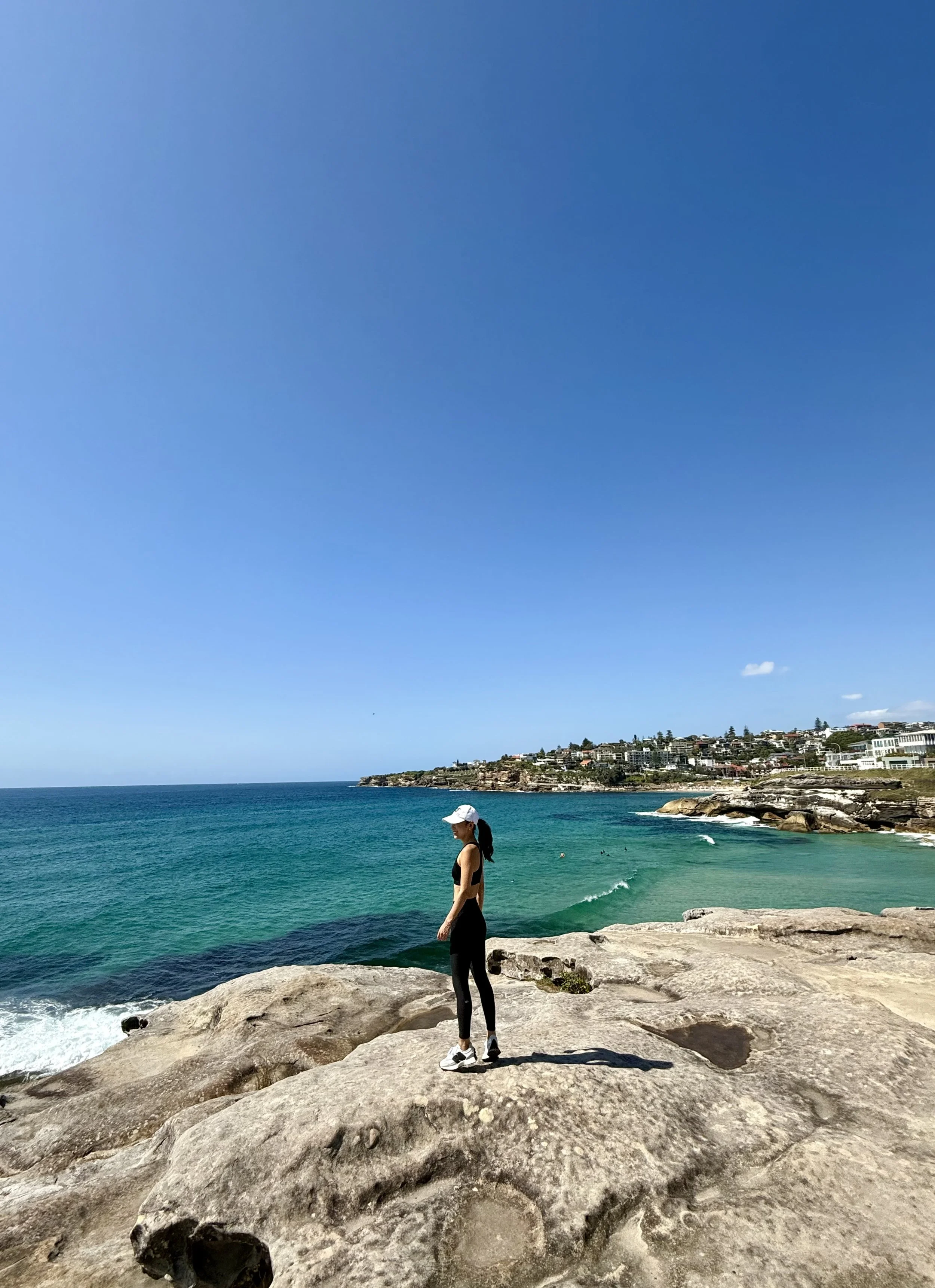 A woman in athletic clothing and a white cap standing on rocky shoreline overlooking the ocean with cliffs and houses in the background under a clear blue sky.