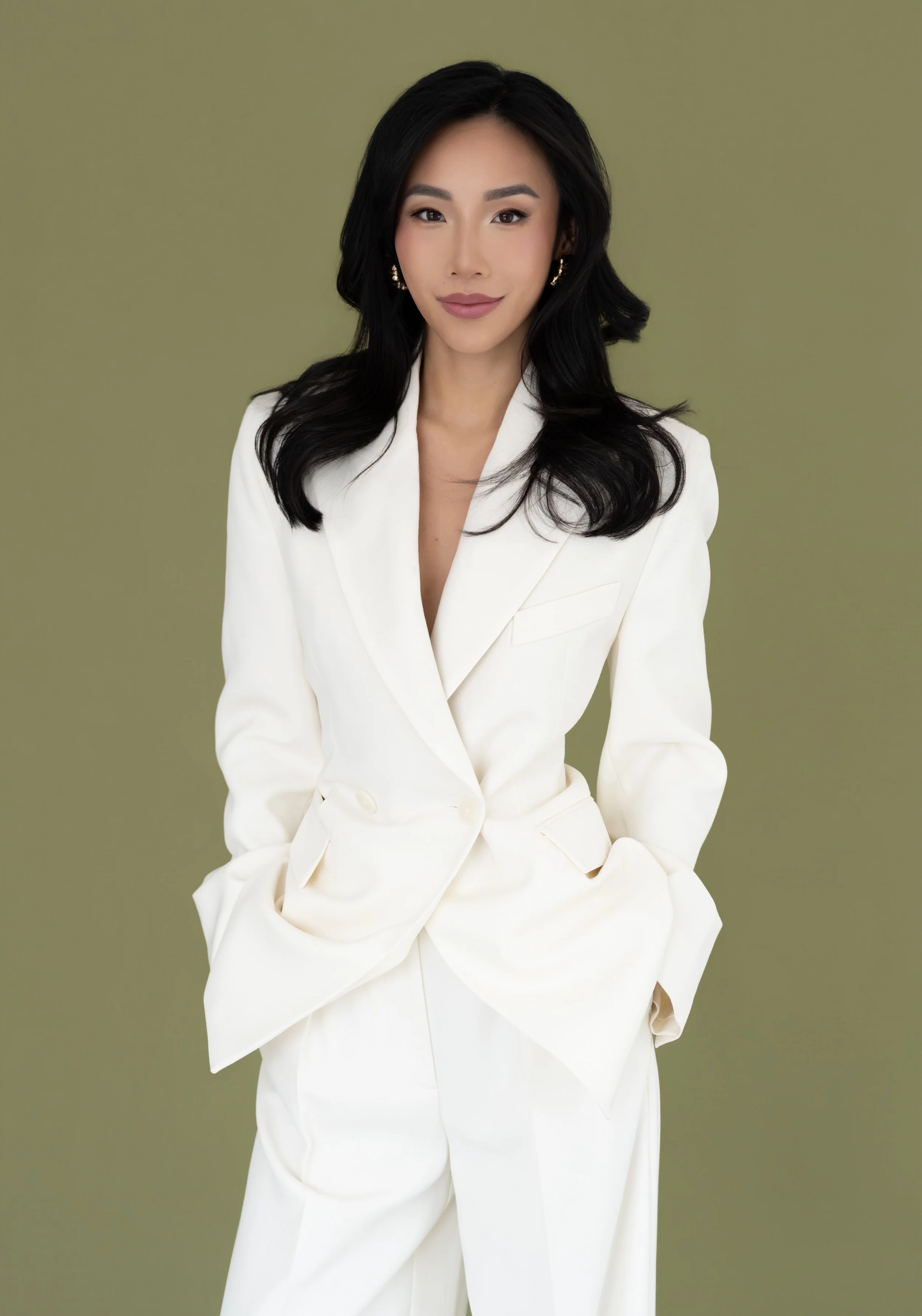 A woman with black wavy hair wearing a white suit, sitting against an olive green background.