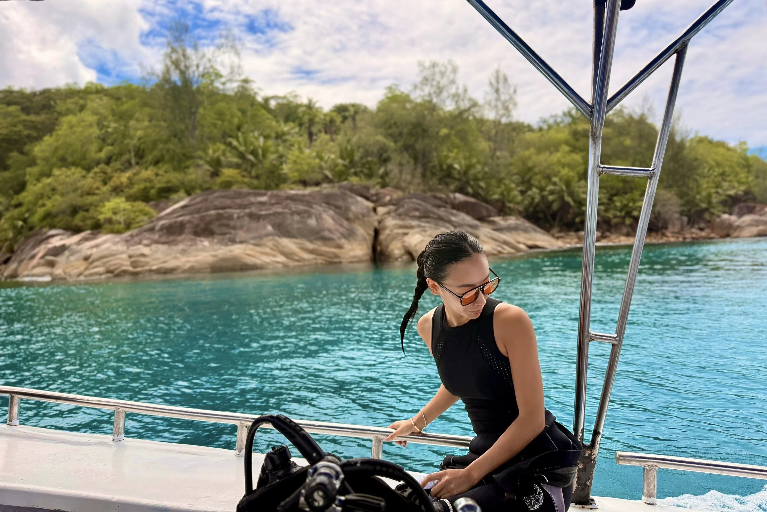 A woman with braided hair and sunglasses sitting on a boat, with a scenic background of water, green trees, rocky shoreline, and partly cloudy sky.