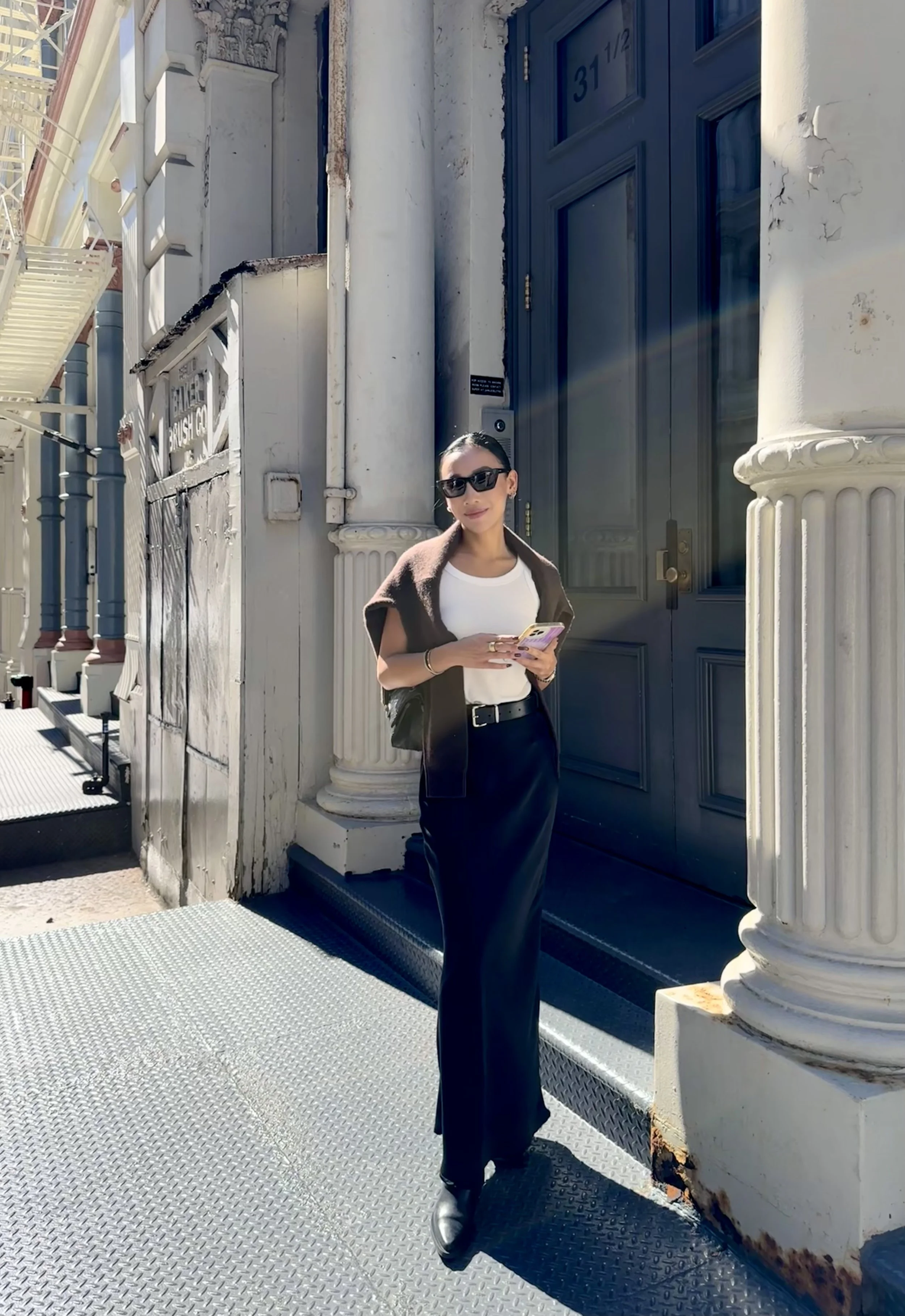 A woman wearing sunglasses, a white shirt, black pants, and a brown jacket draped over her shoulders, standing in front of a building with white columns and a large blue door, looking at her phone.
