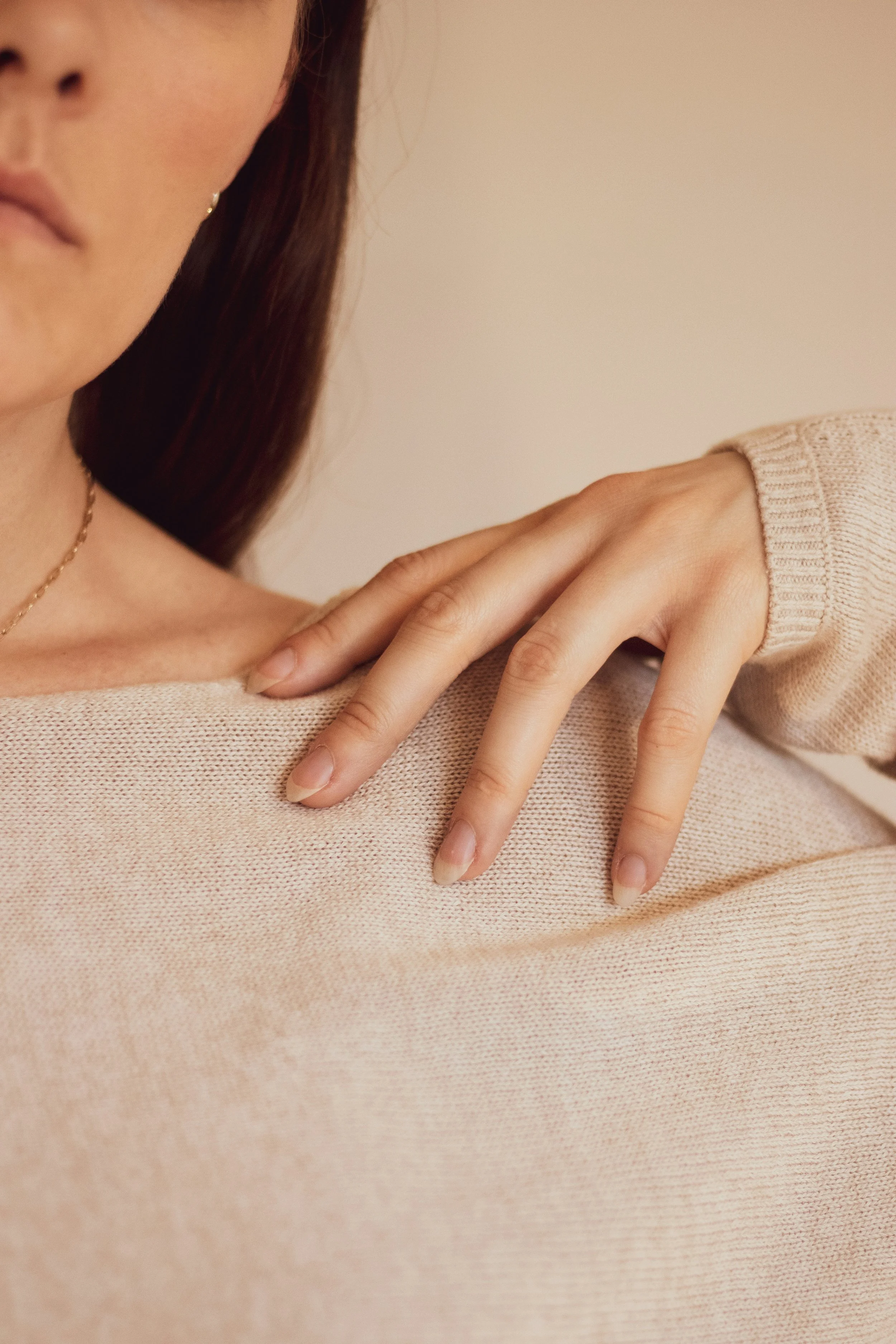 Close-up of a woman with brown hair, partially visible face, wearing a beige sweater, with her hand resting on her shoulder.