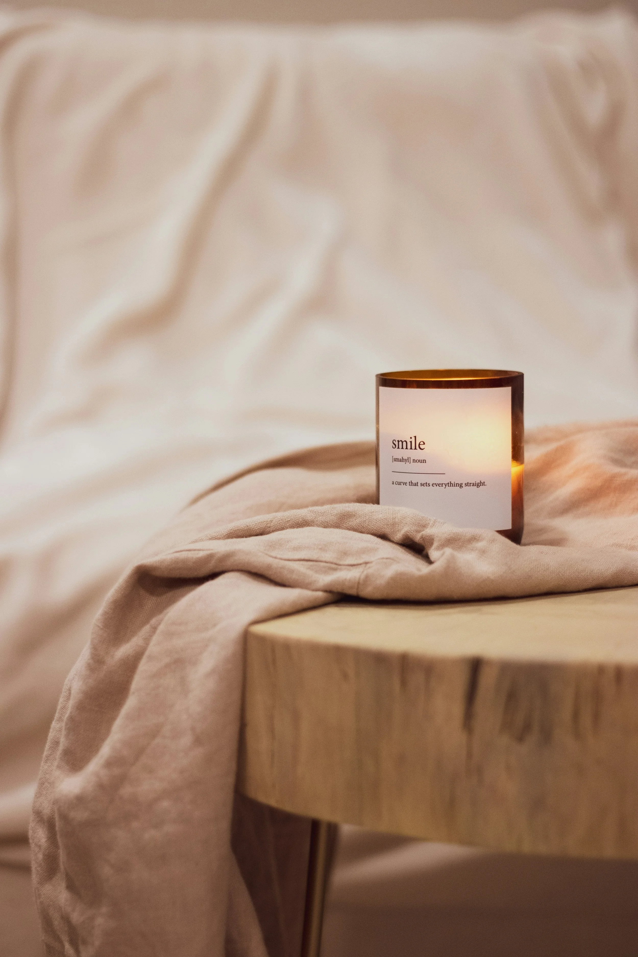 A candle on a wooden table with a warm glow, displaying the word 'smile' and its definition as a phrase, against a soft, beige fabric backdrop.