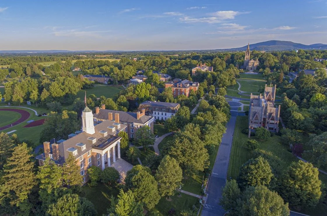 Aerial view of a college campus surrounded by trees, with multiple buildings including a church with a tall spire, open green spaces, athletic track, and distant hills under a partly cloudy sky.