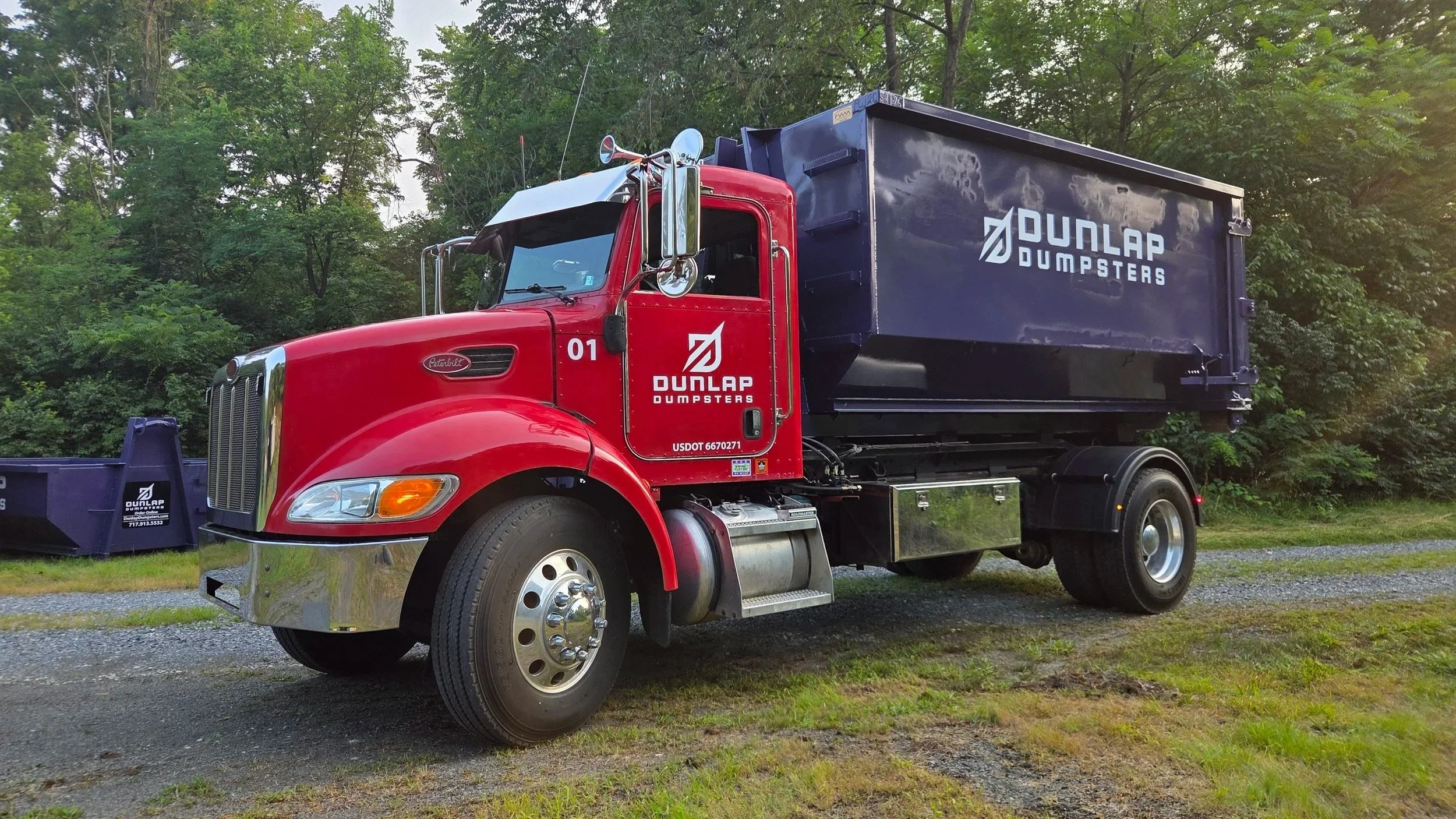 Red and black dumpster truck with 'Dunlap Dumpsters' logo parked on grassy ground near trees.