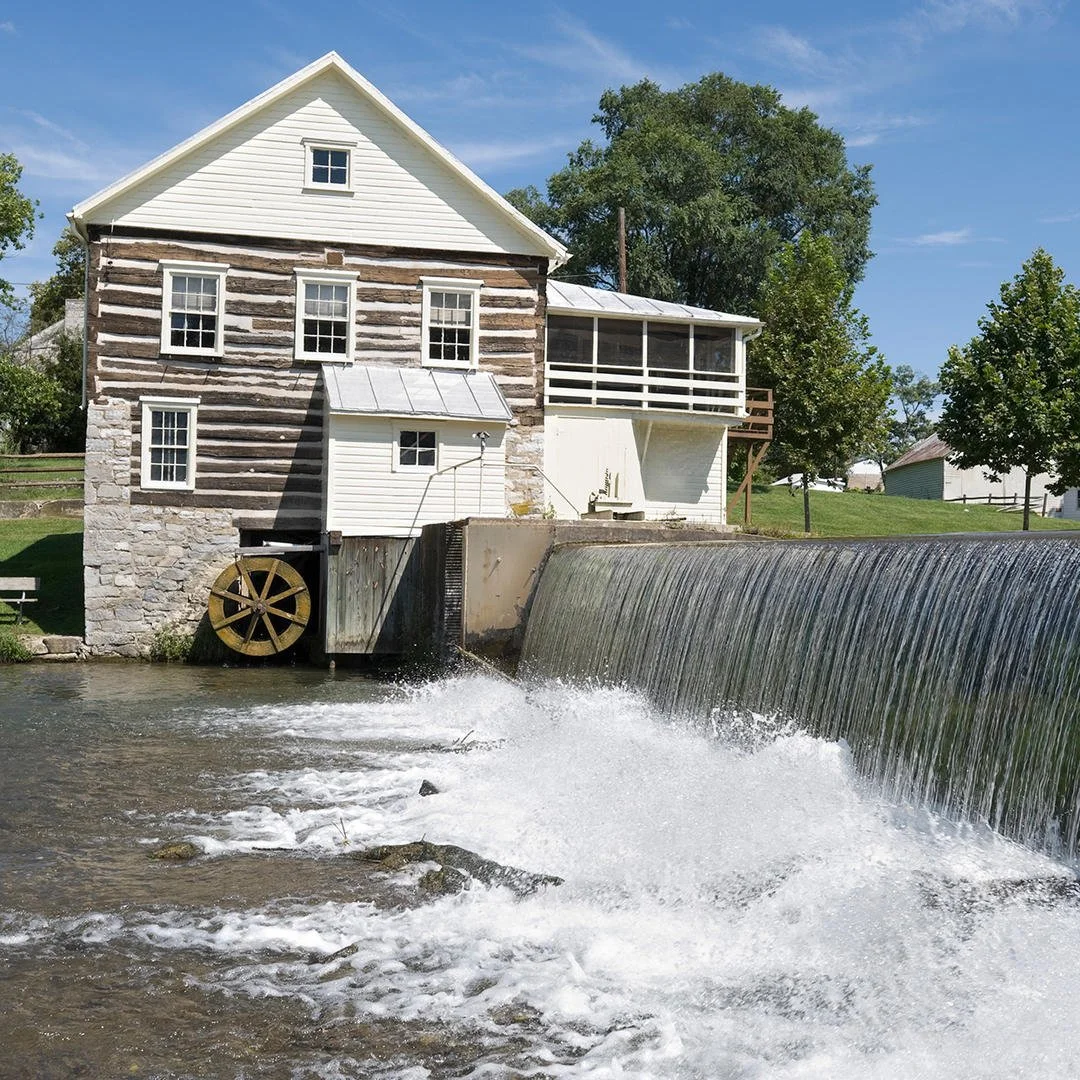 A historic watermill house with a stone and wood exterior, a small upper window, and a water wheel by a dam on a river, surrounded by trees and greenery, under a blue sky.