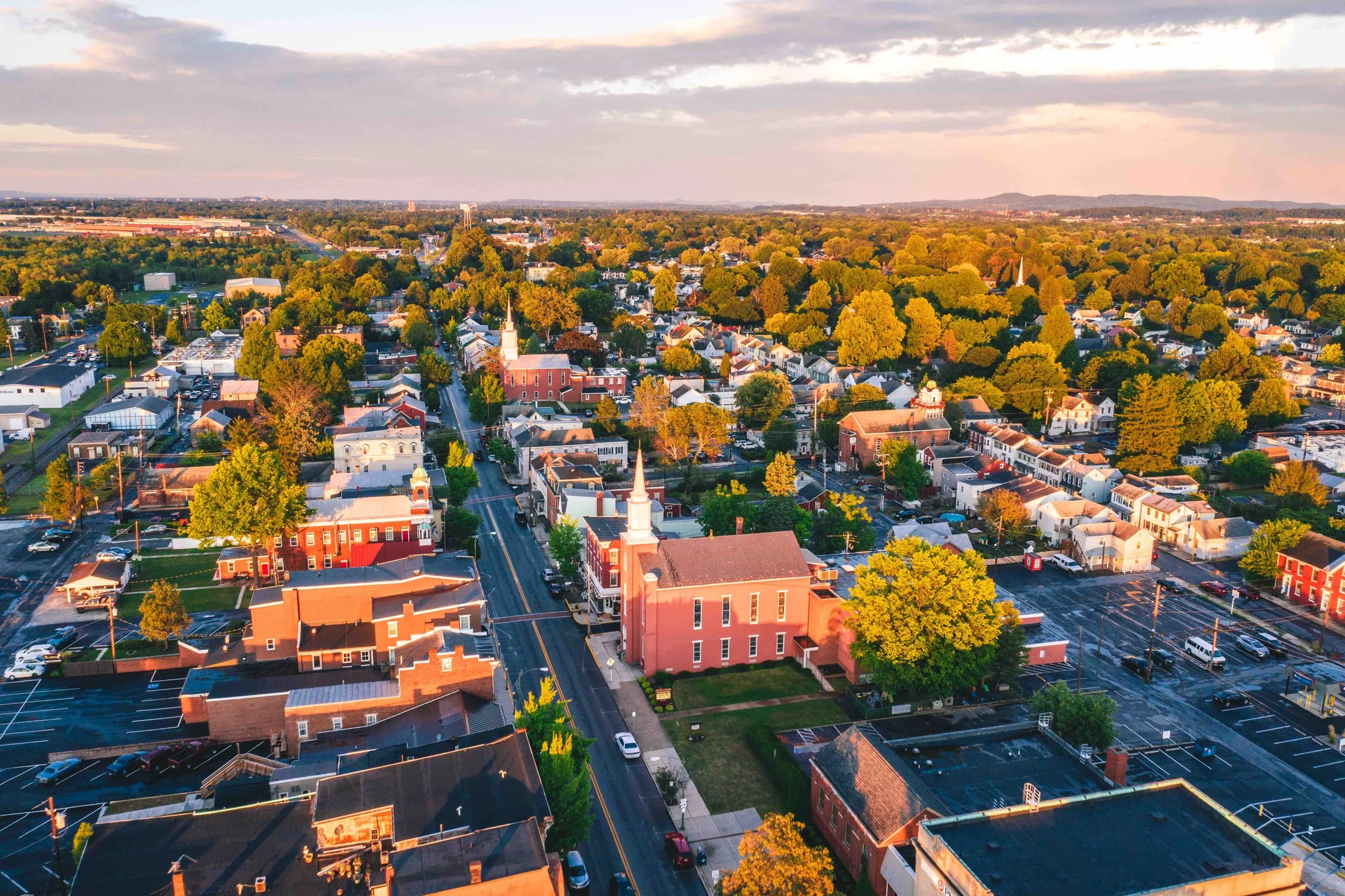 Aerial view of a small town surrounded by many green trees and pastures, with a few church steeples and commercial buildings, under a partly cloudy sky during sunset.