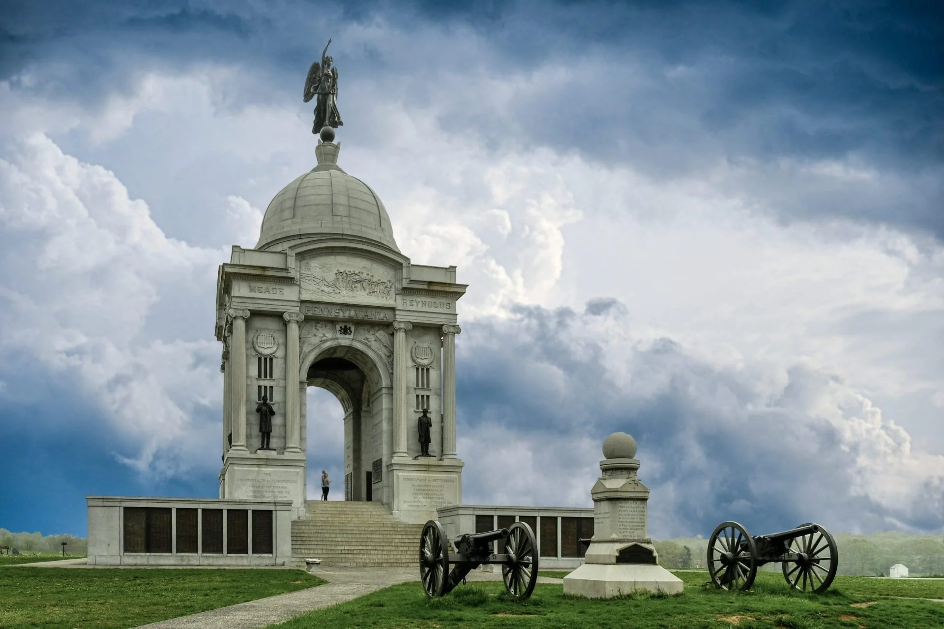 A historical monument with a dome and statues, with cannons in the foreground and a cloudy sky in the background.