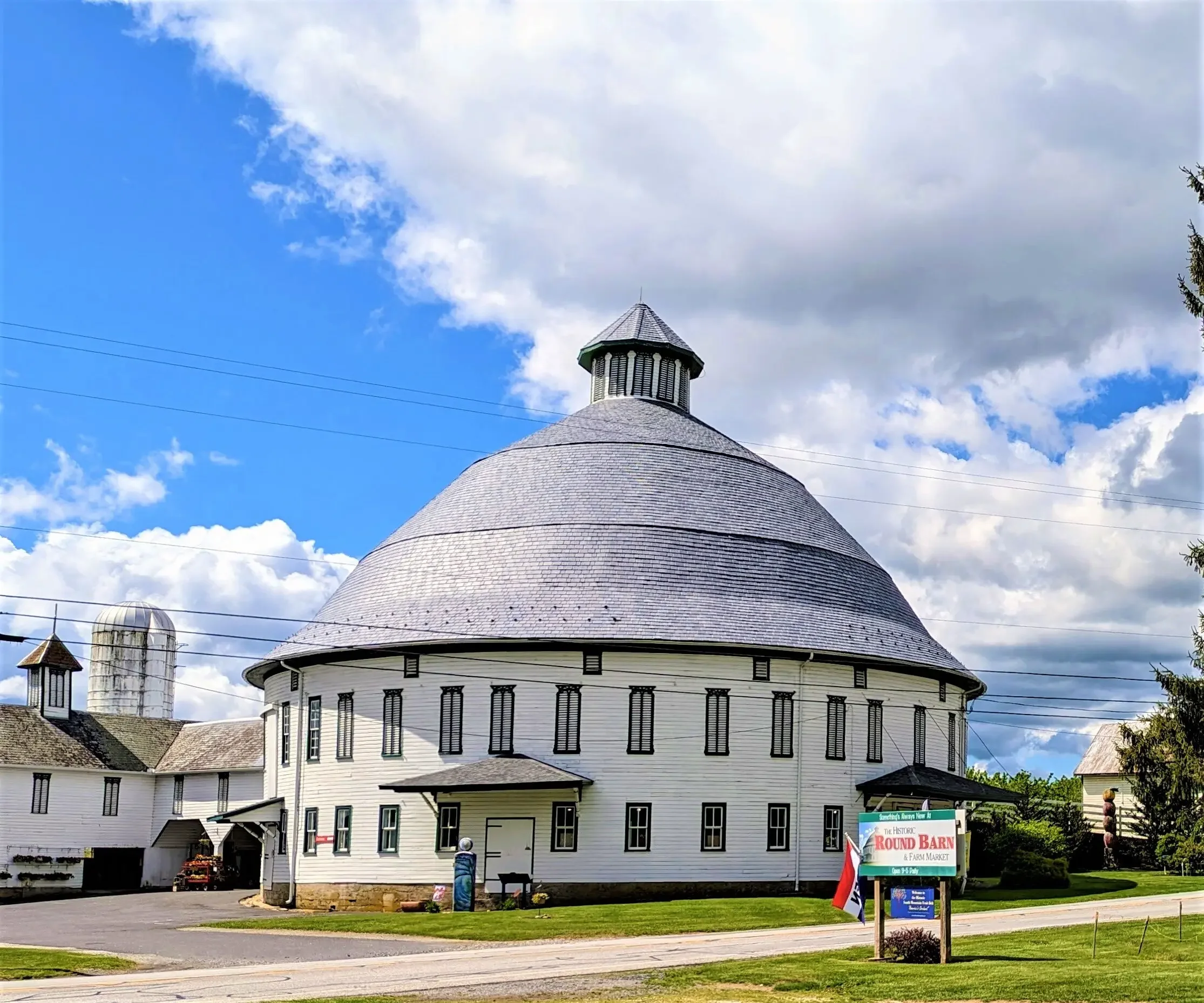 A large white round barn with black trim and small windows, topped with a conical roof and small cupola, located under a partly cloudy blue sky. There is a sign that reads 'The Historic Round Barn & Farm Market' and a flag in front of it.