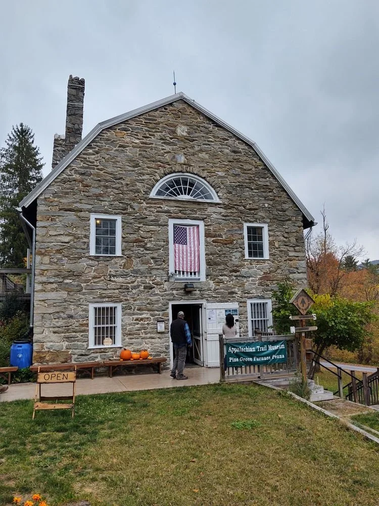 Stone building with an American flag hanging on the front, surrounded by trees and a grassy area, with pumpkins on a bench outside and a sign for the Appalachian Trail Museum.