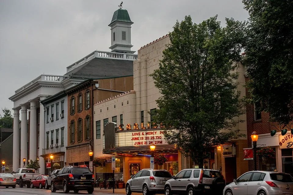 Street view of a small town with parked cars, brick and beige buildings, a marquee sign, and a white church with columns and a green dome in the background.