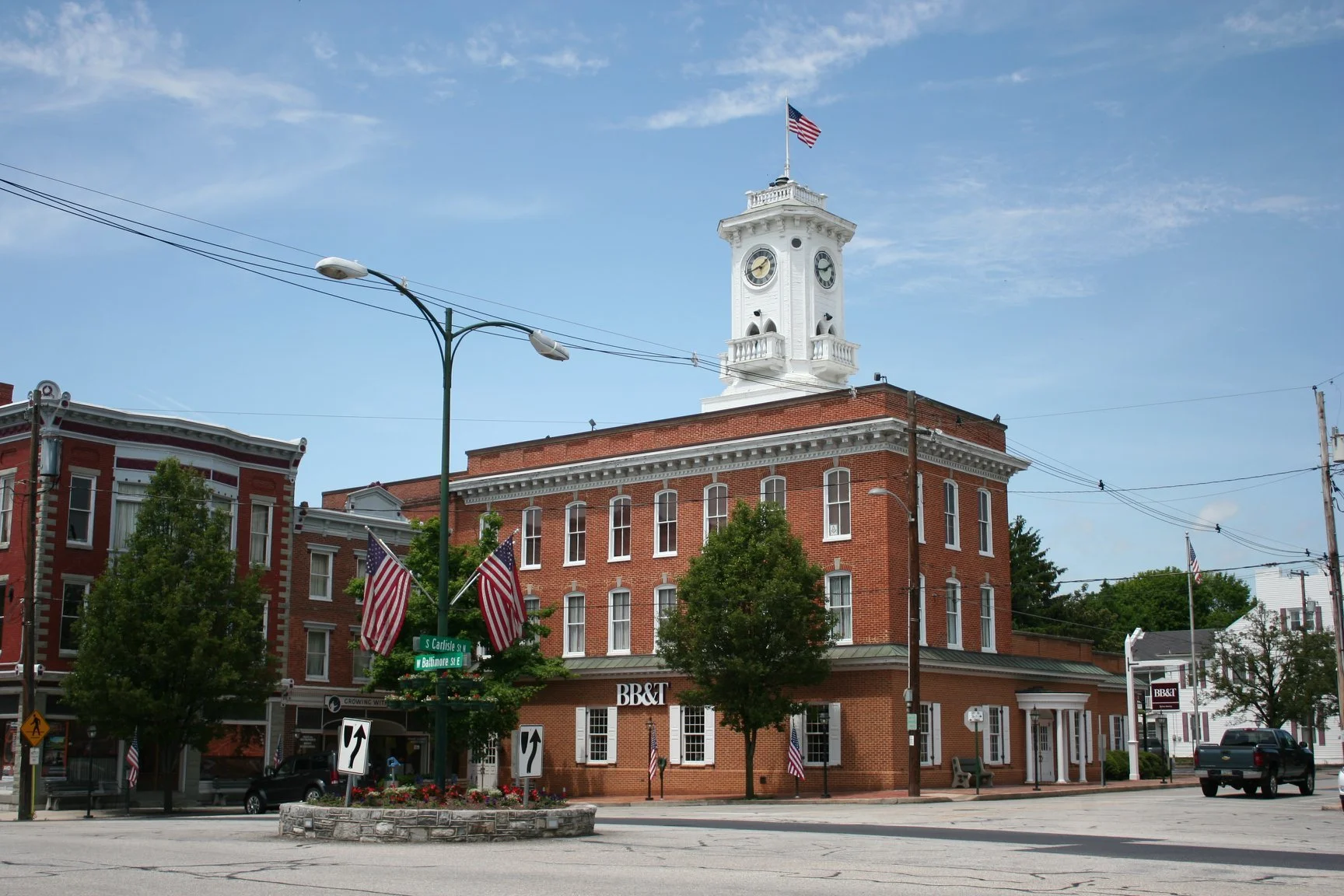 A historic brick building with a clock tower, American flags, and green trees in a small town square under a blue sky.
