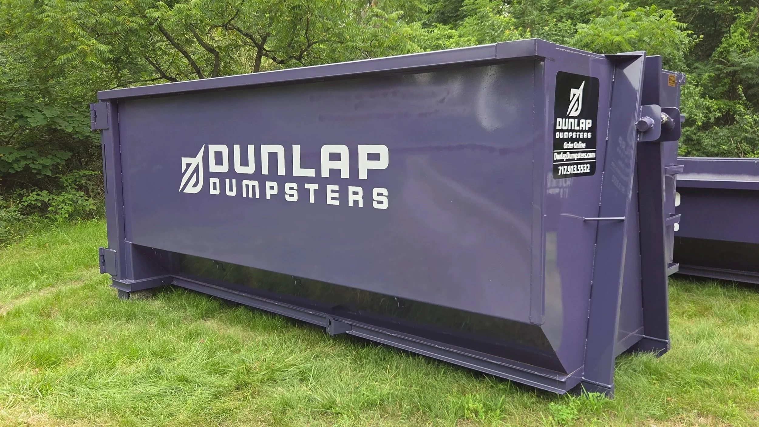 A purple dumpster with white lettering that reads 'Dunlap Dumpsters' on a grassy area, with green trees in the background.