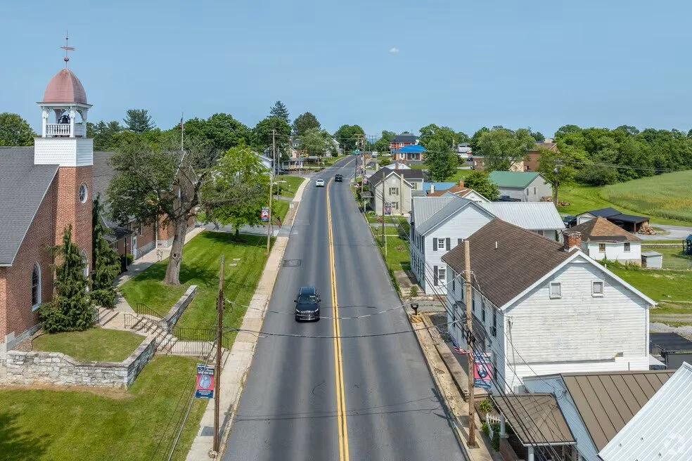 A small town street with cars, residential houses, and a church with a bell tower, surrounded by green trees and fields.