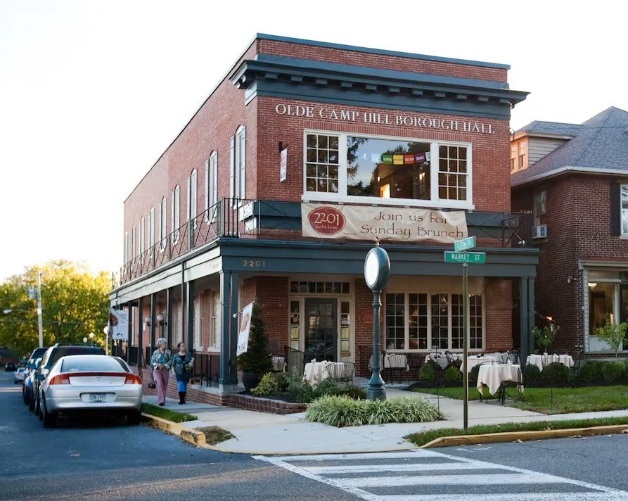 A two-story brick building with the sign 'Olde Camp Hill Borough Hall' and a banner inviting people to a Sunday brunch. The building has large front windows and outdoor dining tables with white tablecloths on the patio. A street sign indicates the location as Market Street, and there are parked cars and two people walking on the sidewalk.