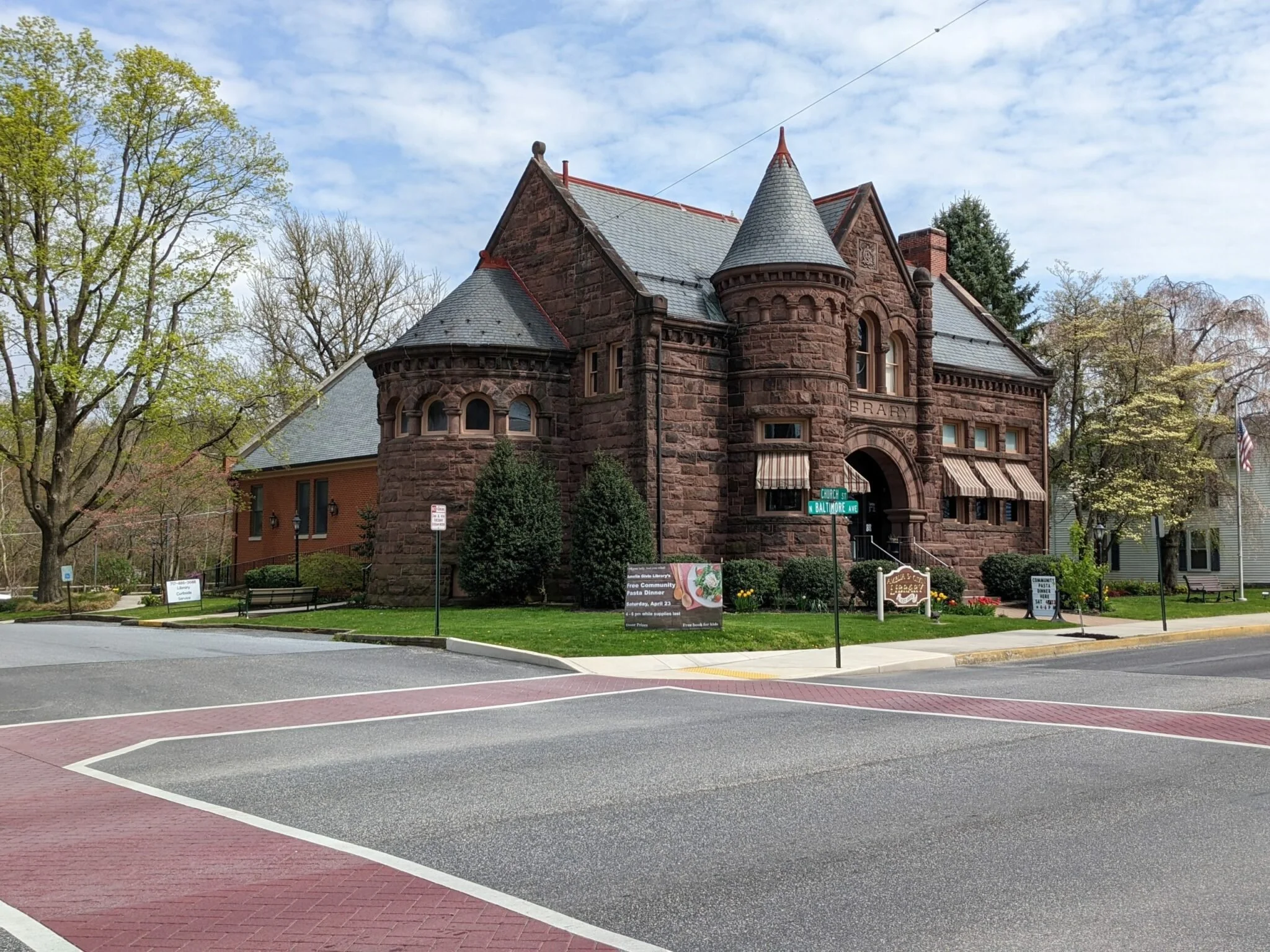 A historic stone building with a round turret, triangular roof, arched windows, and a sign that says 'BRARY.' There are street signs indicating the intersection of Church Street and North Baltimore Avenue, surrounded by trees and a small park area with signs, benches, and flower beds. The sky is partly cloudy.