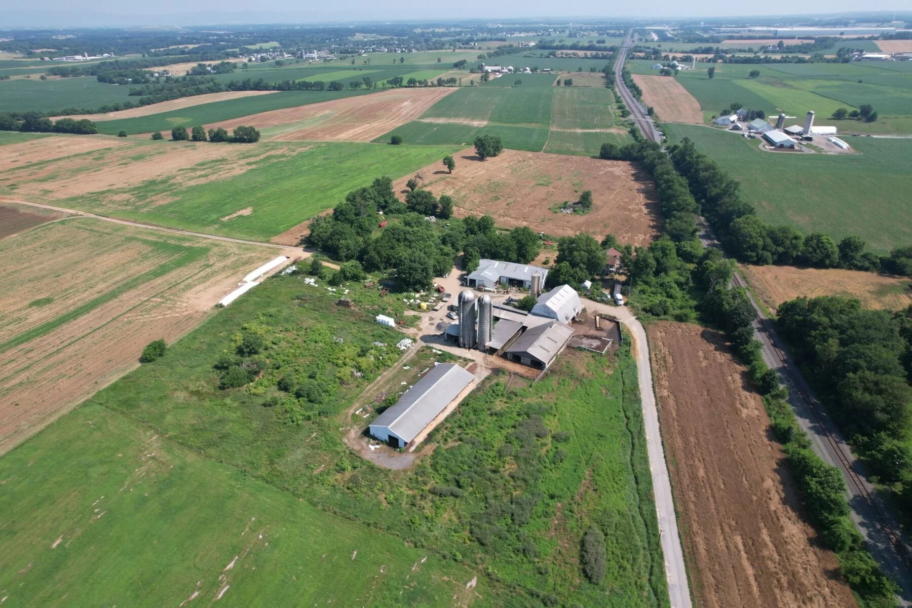 Aerial view of farmland showing fields, trees, farm buildings, silos, and a railway track running through the landscape.
