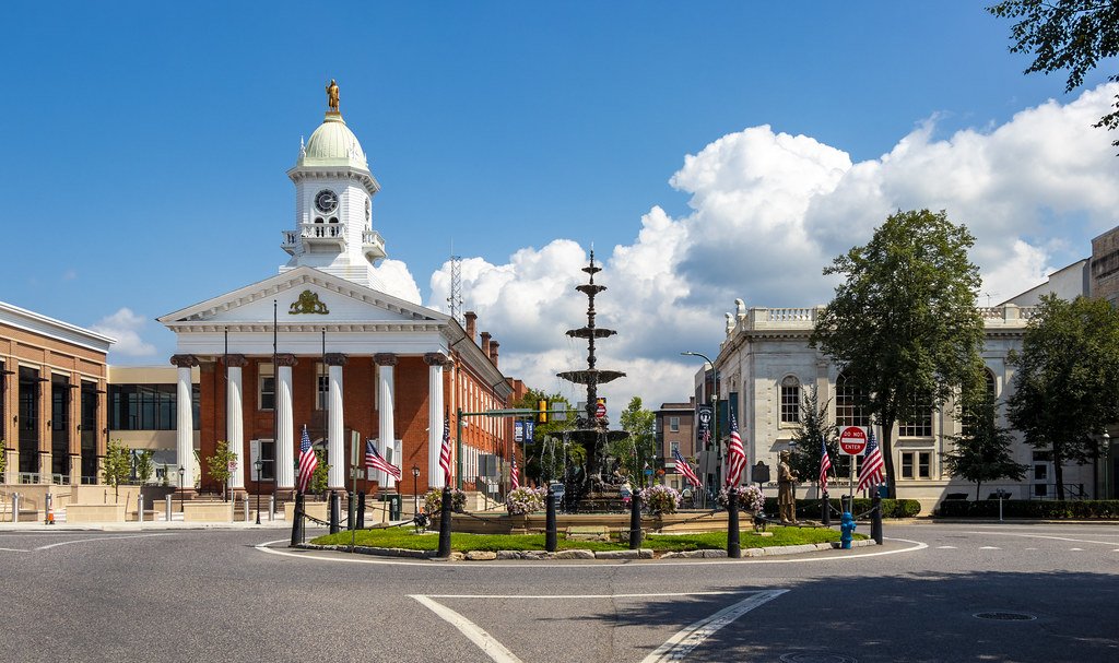 A city square with a fountain in the center, decorated with American flags, in front of a historic building with a green dome and statue on top, under a bright blue sky with clouds.