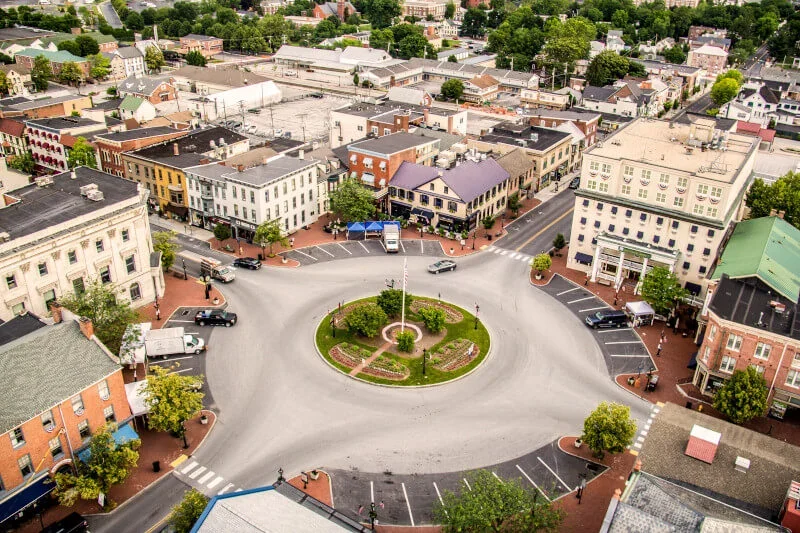 Aerial view of a small town's roundabout with a green central island and parking spaces around it, surrounded by various buildings and trees.